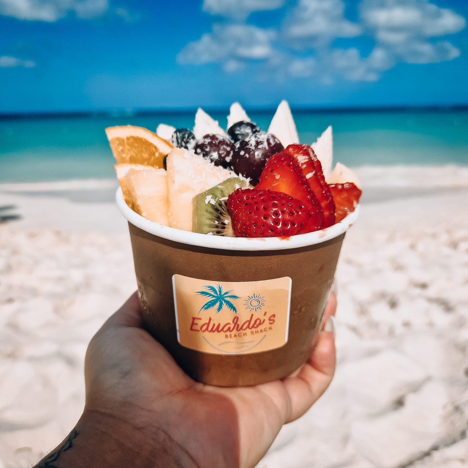 Hand holding Eduardo's tropical fruit cup with strawberries, kiwi, and coconut on a sunny Caribbean beach.