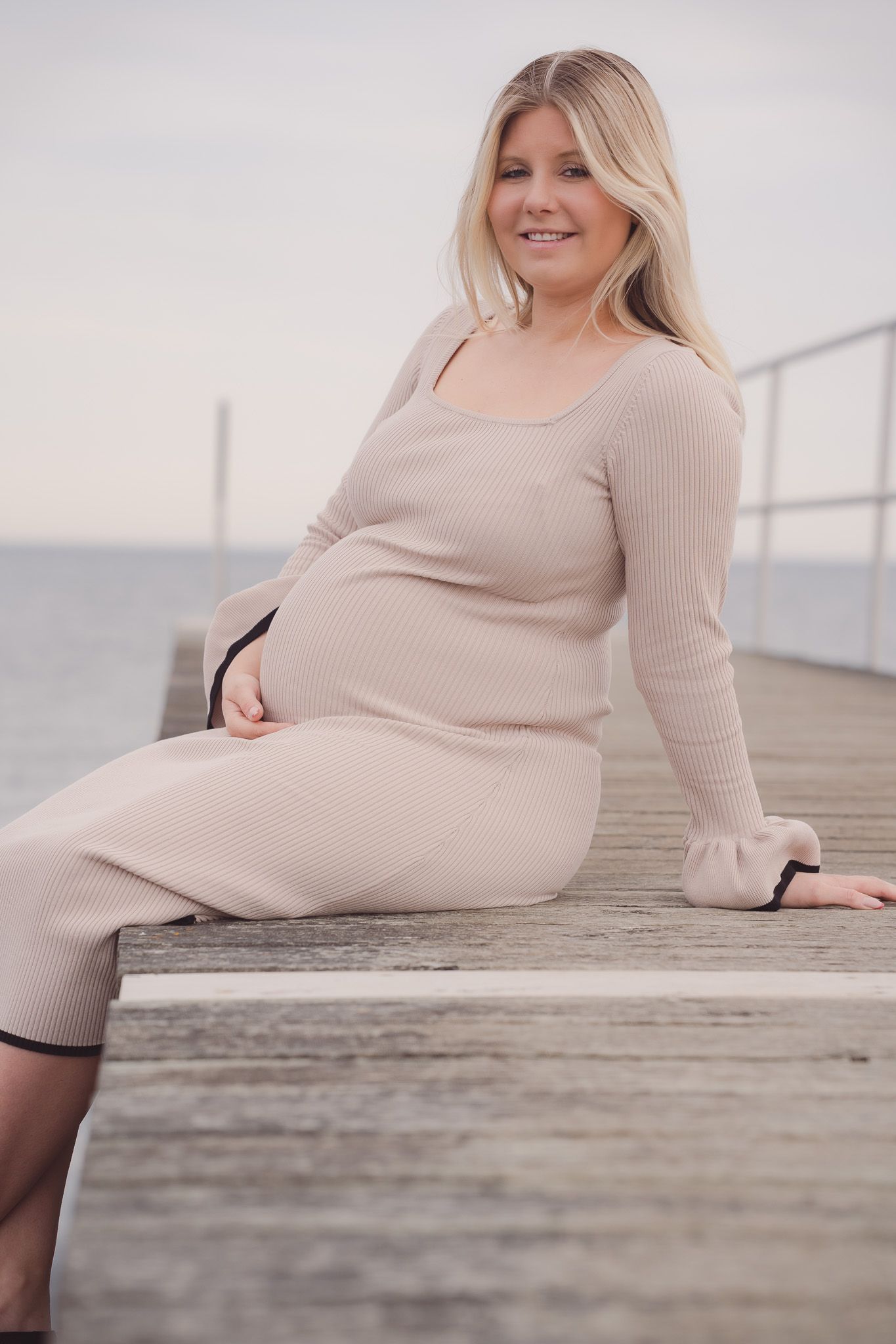 Smiling pregnant woman in beige ribbed dress sitting on wooden pier by the ocean.