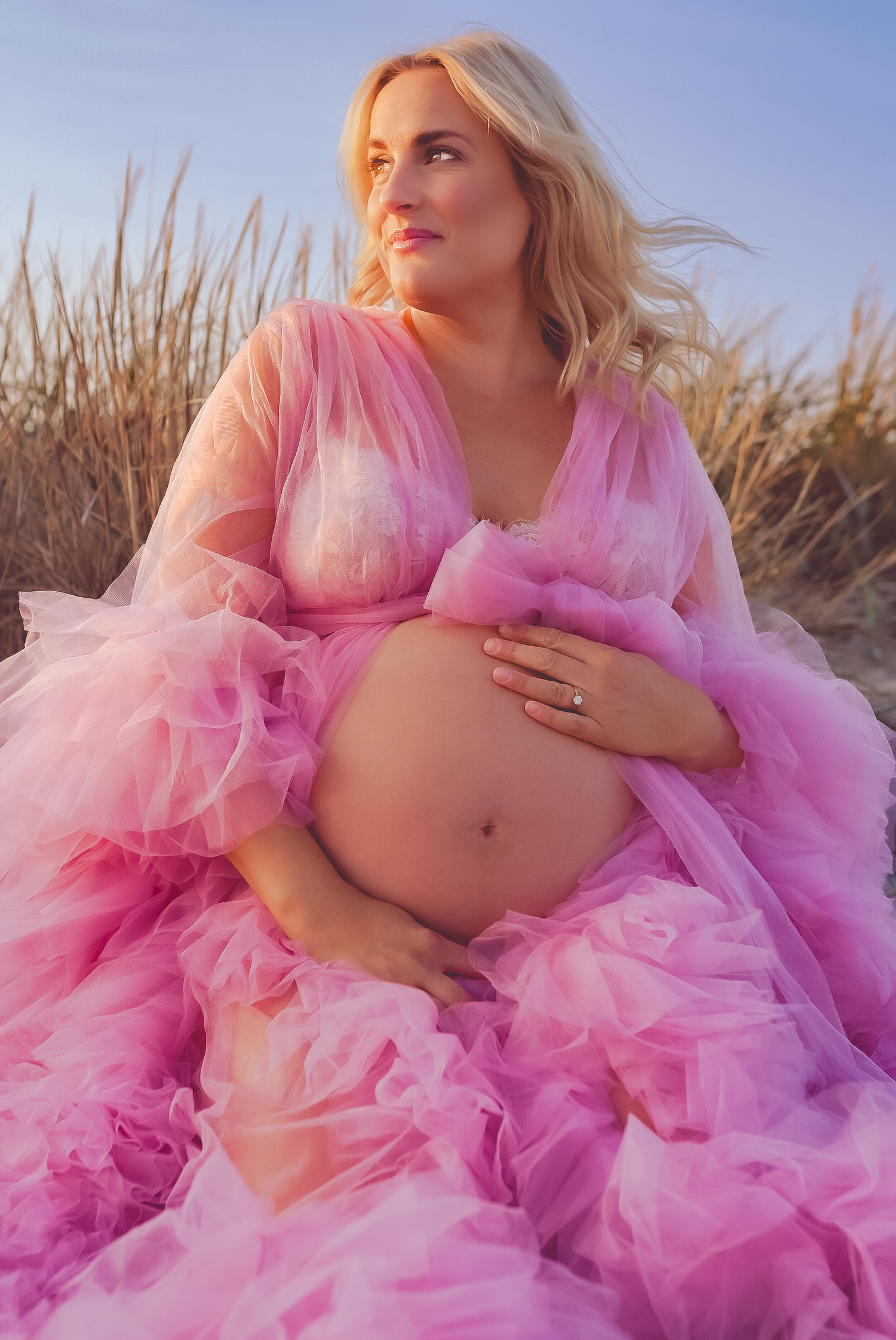 Gravidfotografering på strand med rosa tyllklänning mot molnig himmel.