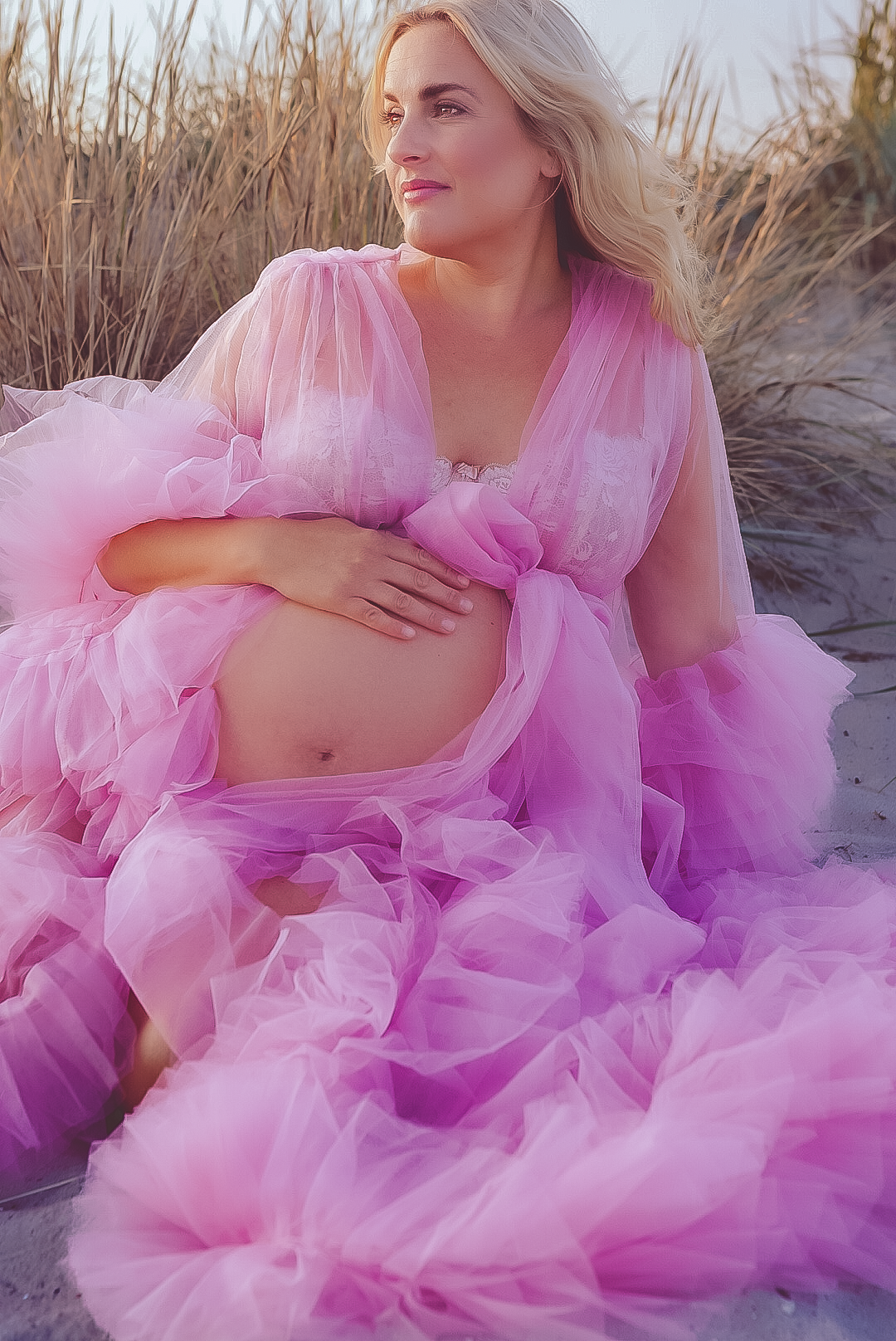 Gravidfotografering på strand med flödande rosa tyllklänning mot högt gräs..