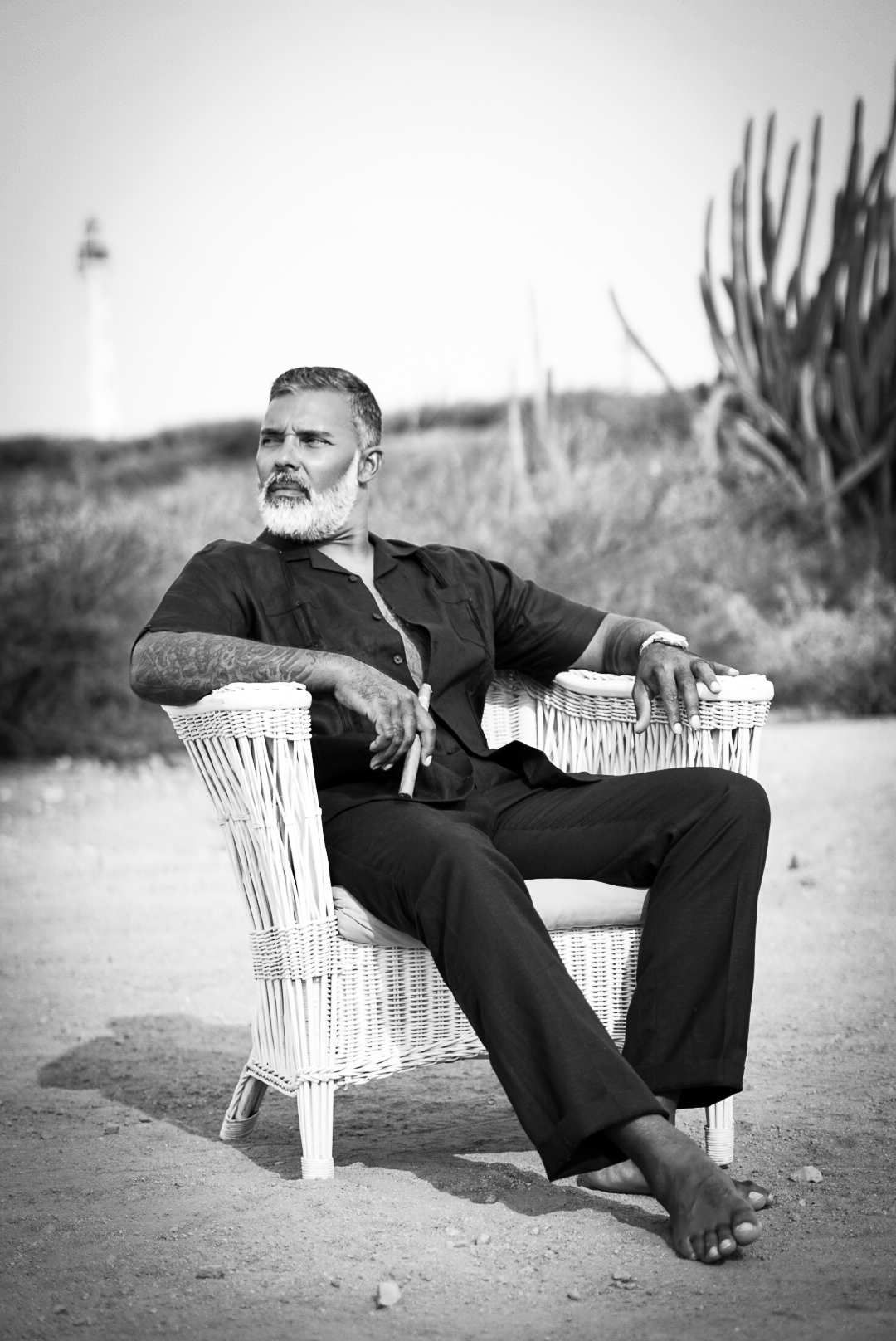 Black and white photo of a bearded man sitting in a wicker chair outdoors near a lighthouse.