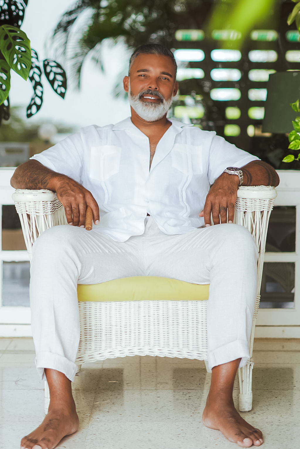 Middle-aged man with gray beard in white outfit sitting relaxed in wicker chair on tropical patio.