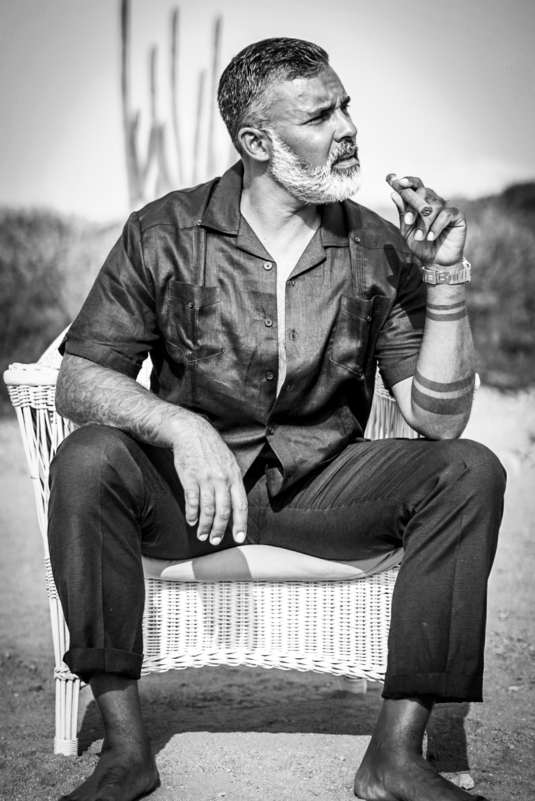 Black and white portrait of a bearded middle-aged man sitting barefoot in a wicker chair outdoors.