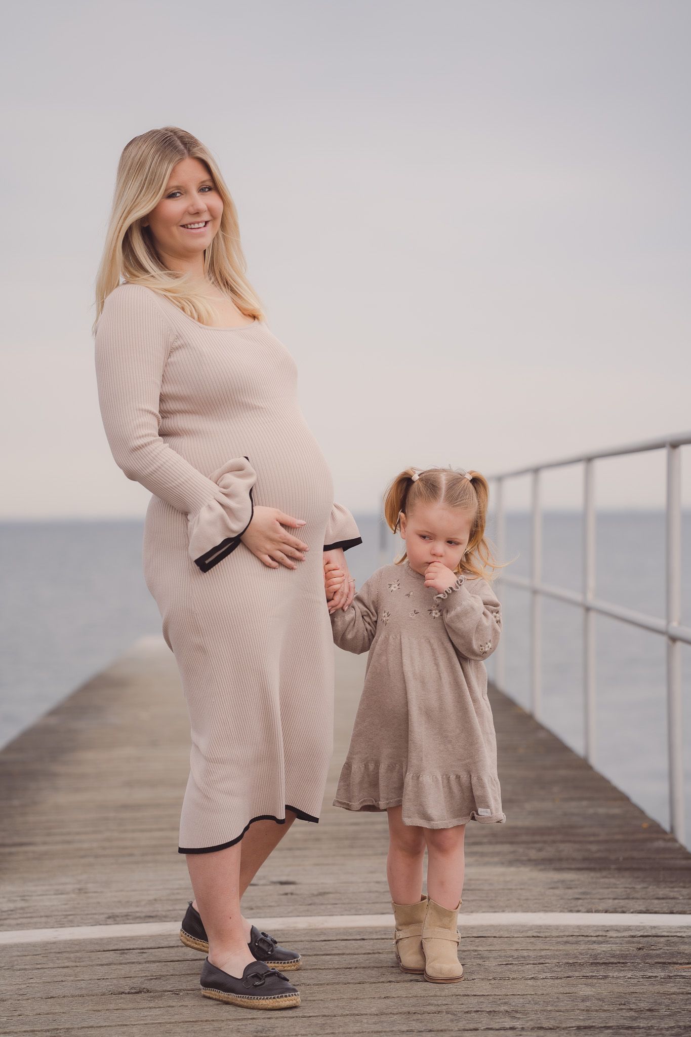 Pregnant woman in beige dress smiles on pier with young daughter in gray dress by the ocean.