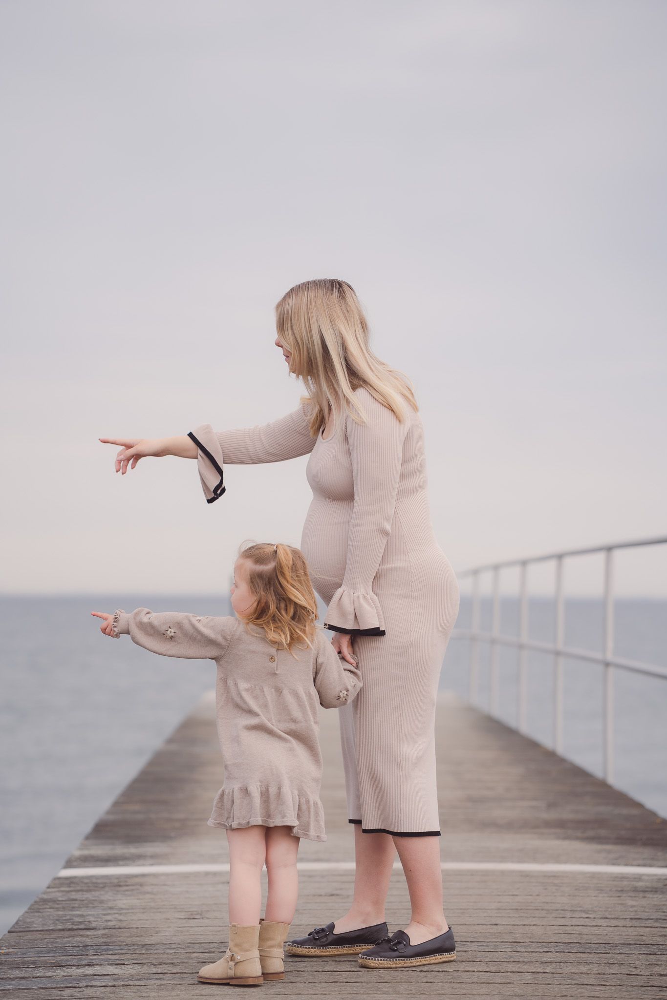 Pregnant woman and young girl pointing together on a seaside pier, both wearing matching beige dresses.