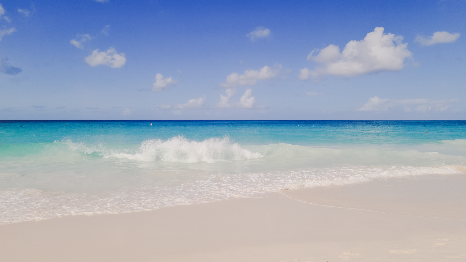 Turquoise waves gently washing onto a pristine white sandy beach under a bright blue sky with scattered clouds.