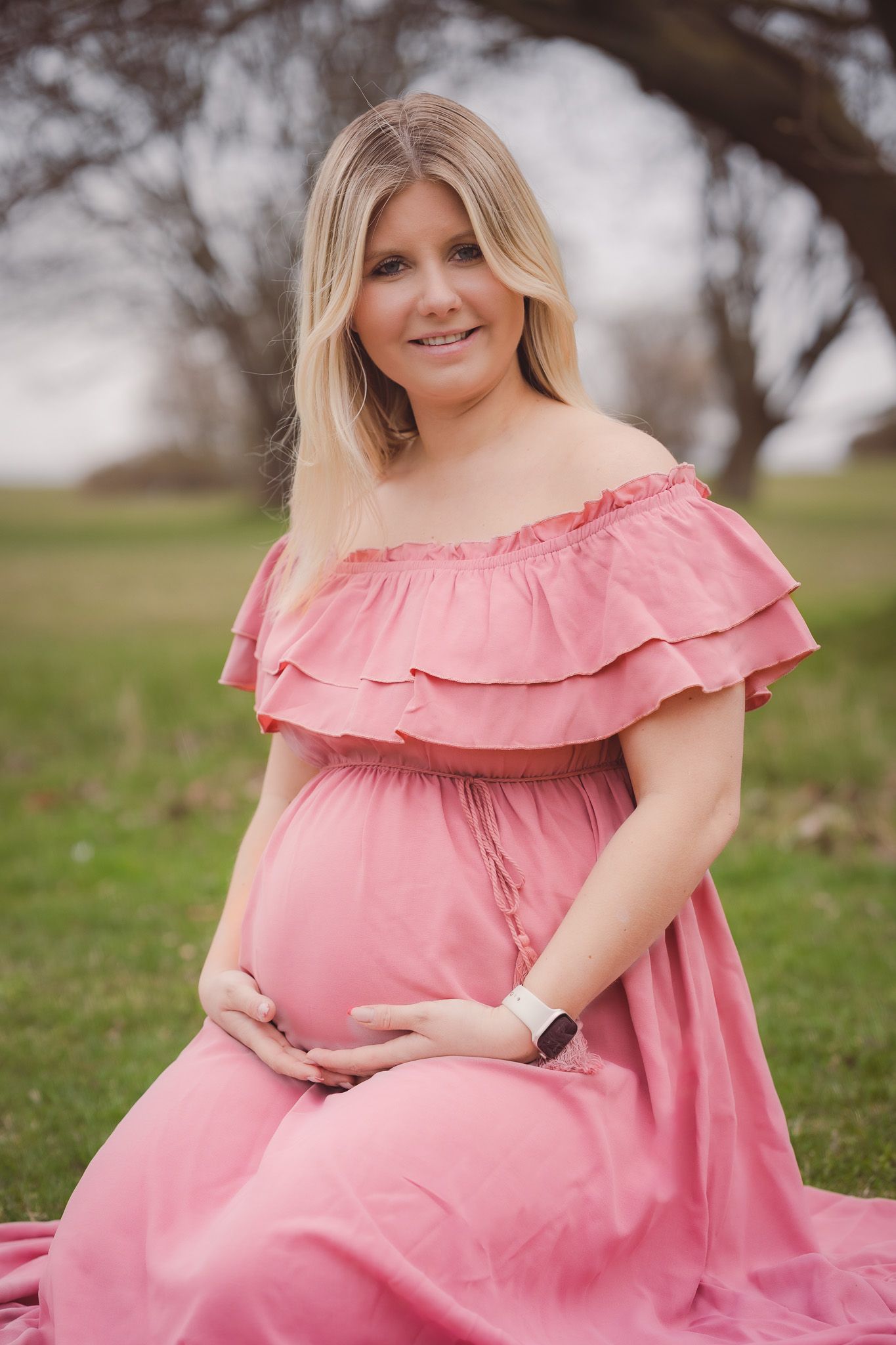 Pregnant woman in pink off-shoulder dress sitting outdoors, cradling baby bump, with green grass background.