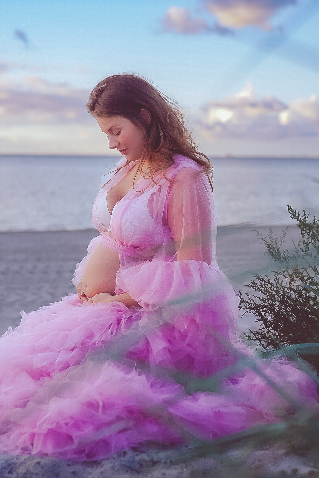 Gravid kvinna i flödande rosa tyllklänning poserar graciöst på stranden vid solnedgång.