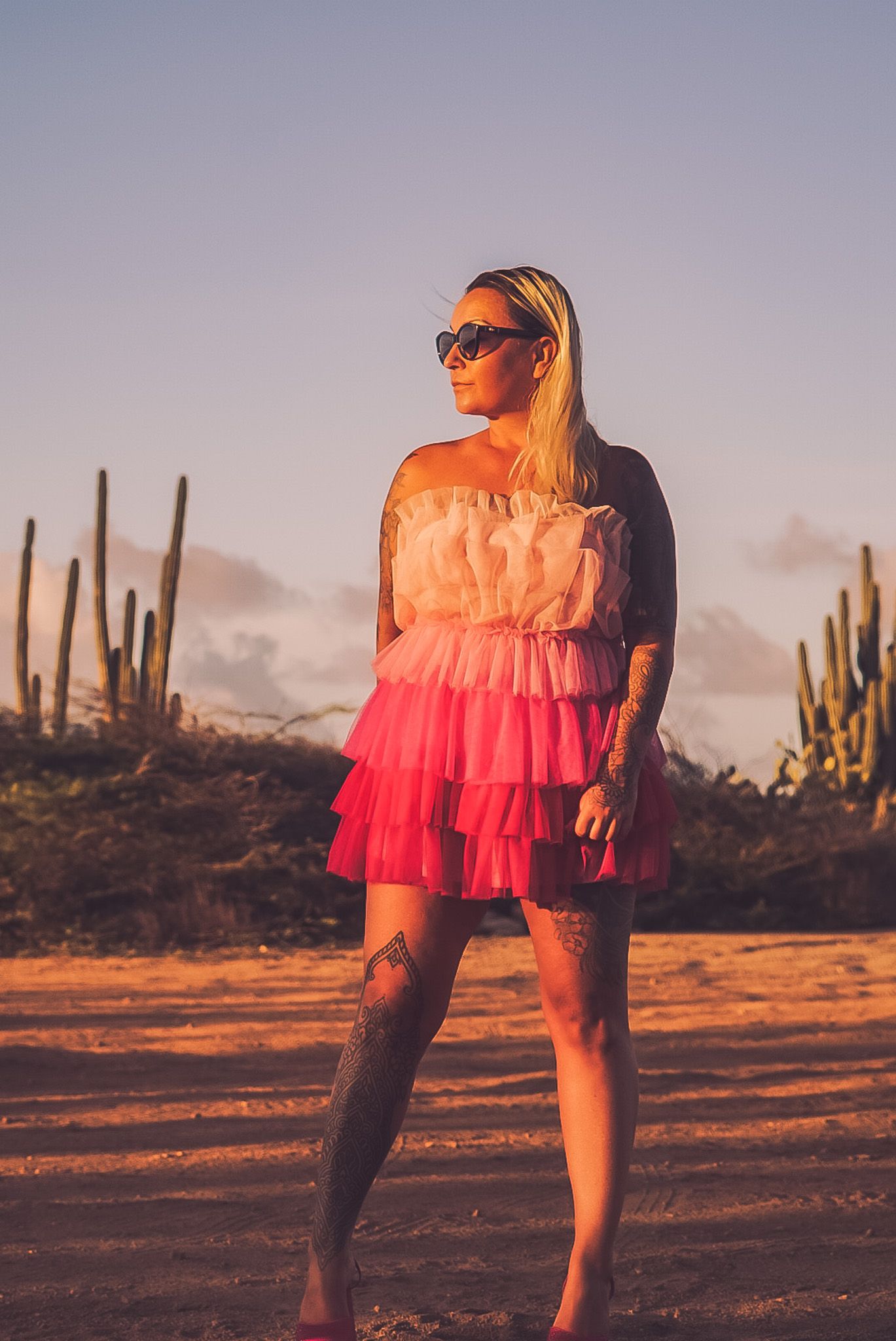 Blonde woman in pink ruffled strapless dress and sunglasses poses in desert landscape with cacti at golden hour.