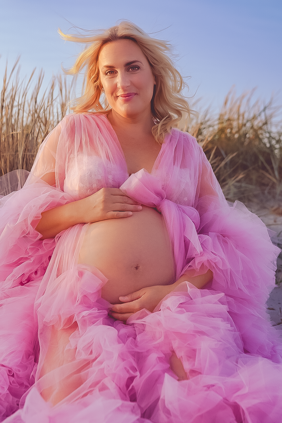 Blivande mamma i rosa tyllklänning poserar på sandstrand vid solnedgång.