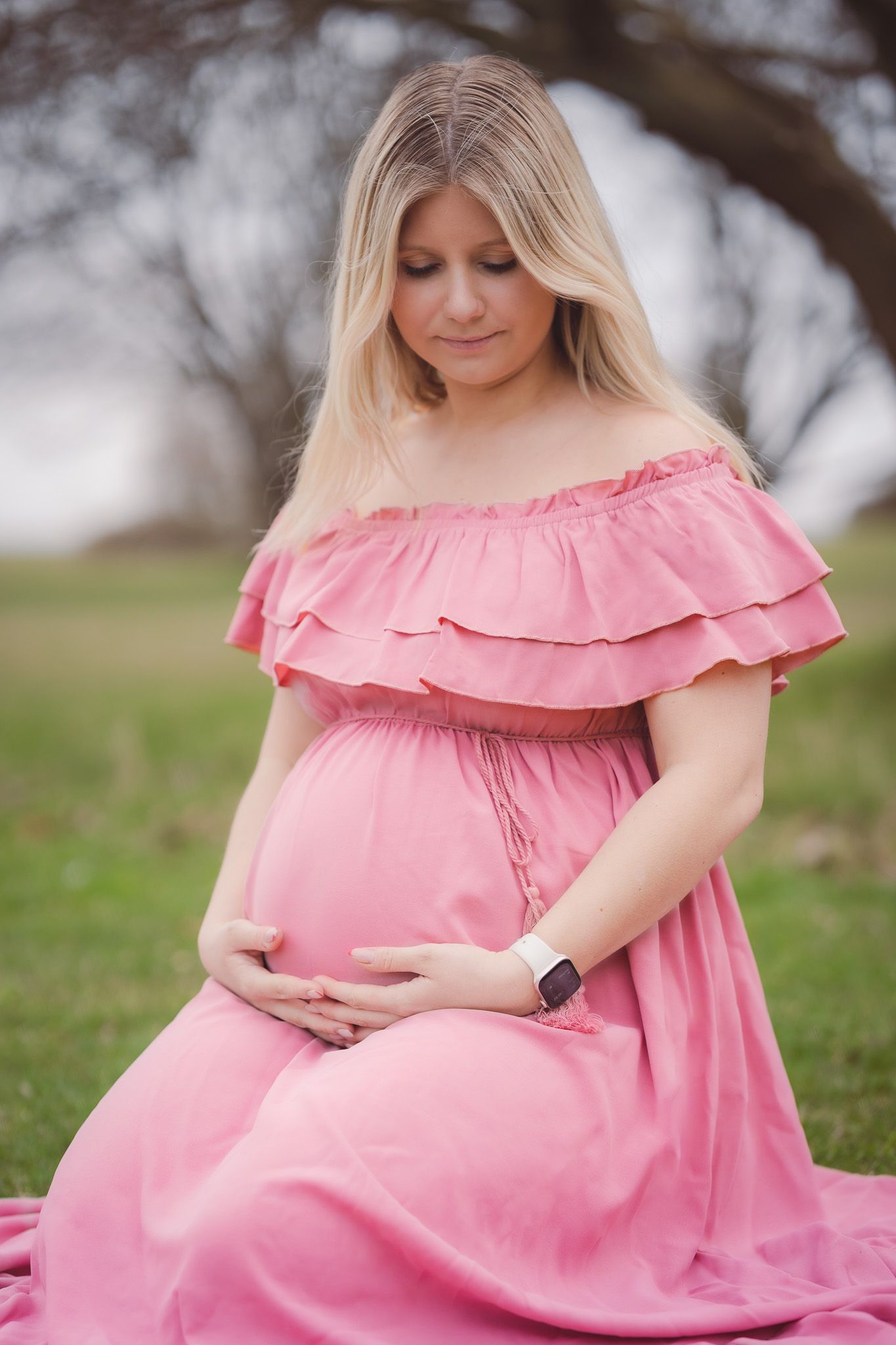 Pregnant woman in pink off-shoulder dress cradling baby bump outdoors in a serene park setting.