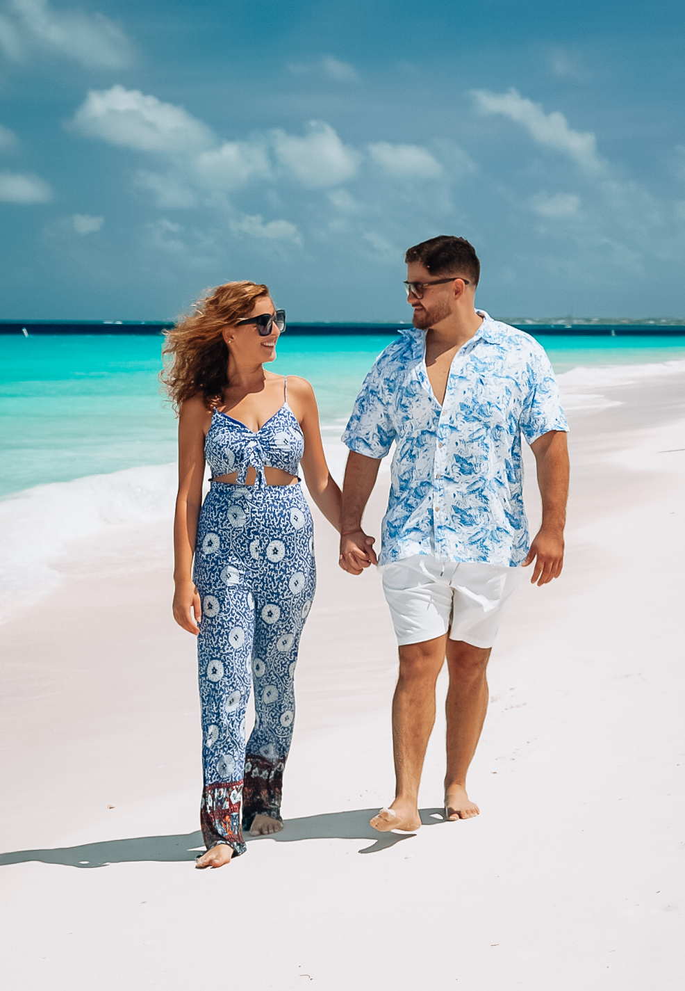 Couple holding hands walking on a white sandy beach, wearing matching blue outfits with turquoise ocean in the background.