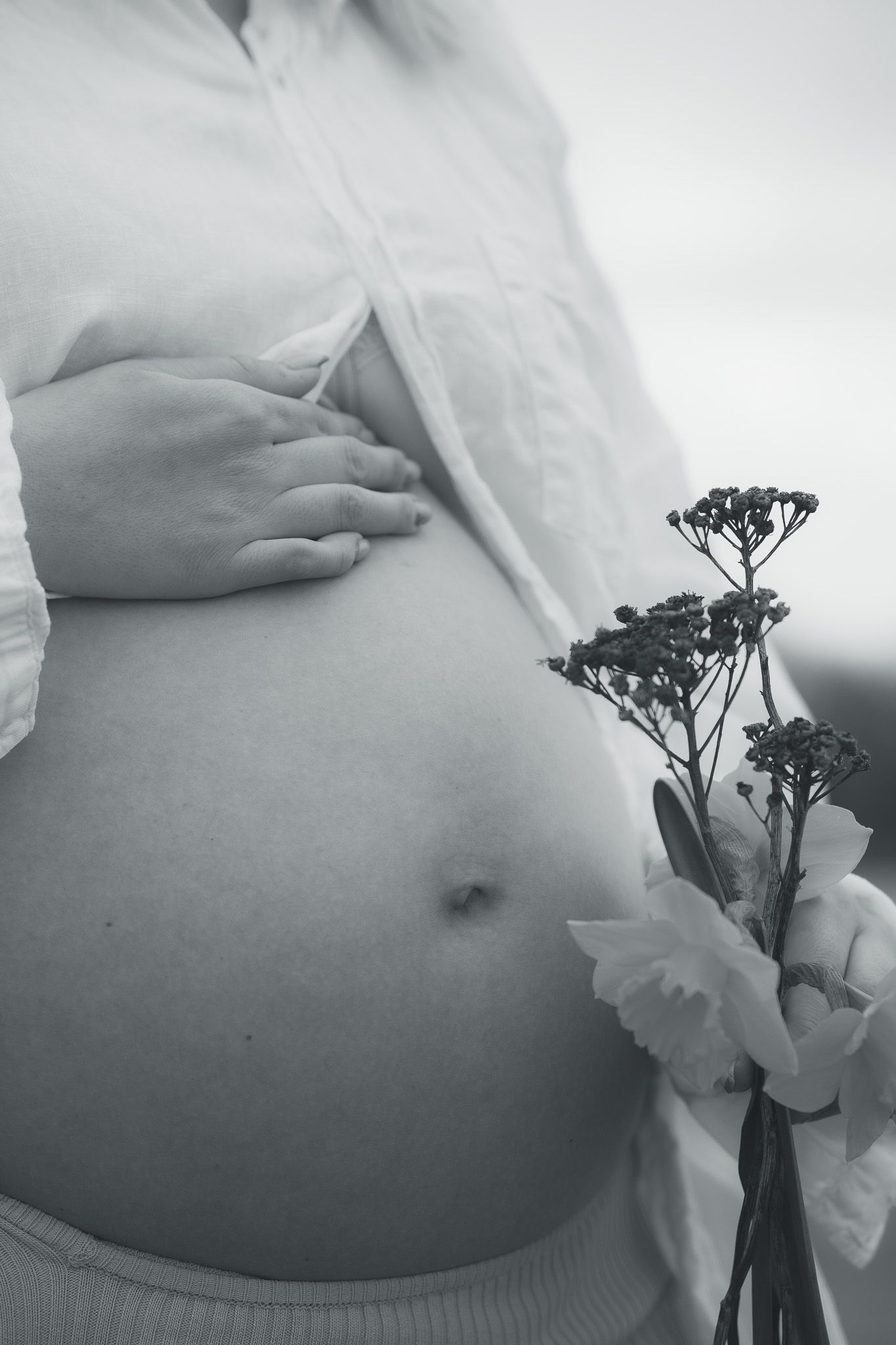 Black and white photo of pregnant woman holding flowers, showing bare baby bump with hand resting gently on stomach.