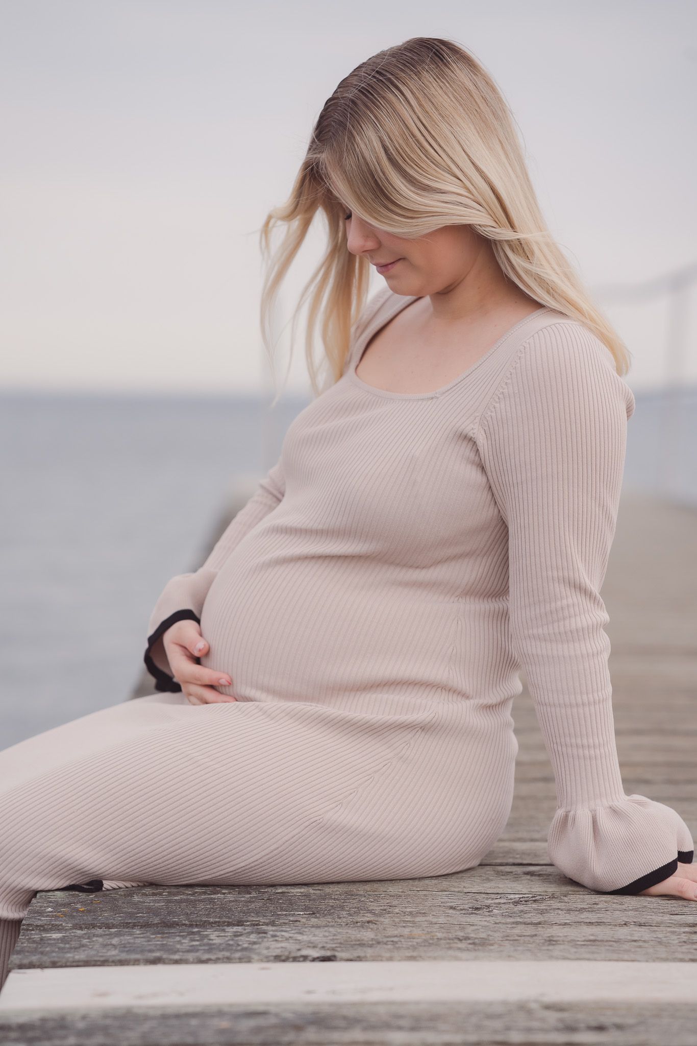 Pregnant woman in beige ribbed dress sitting on wooden pier, cradling baby bump, with ocean in background.