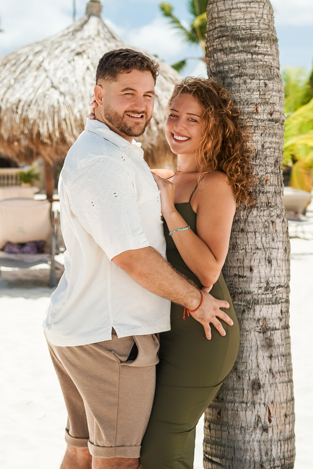 Smiling couple embracing by a palm tree on a sunny tropical beach, woman in green dress, man in white shirt and khaki shorts.