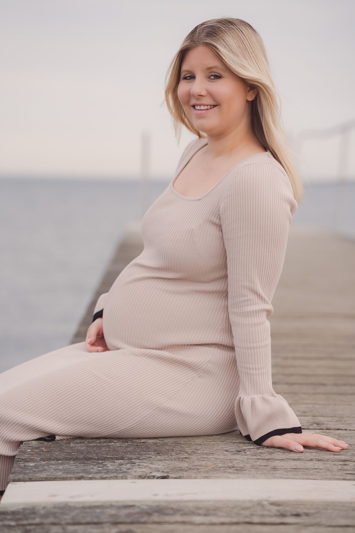 Smiling pregnant woman in beige ribbed dress sitting on wooden pier by the water.