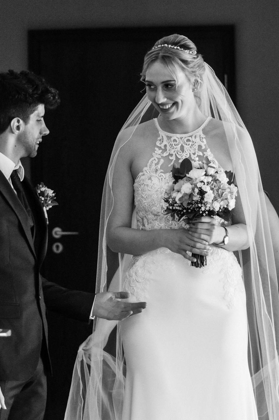 Black & white photo of a smiling bride in lace gown and veil holding bouquet, facing groom in suit at wedding ceremony. Carmela Kolb