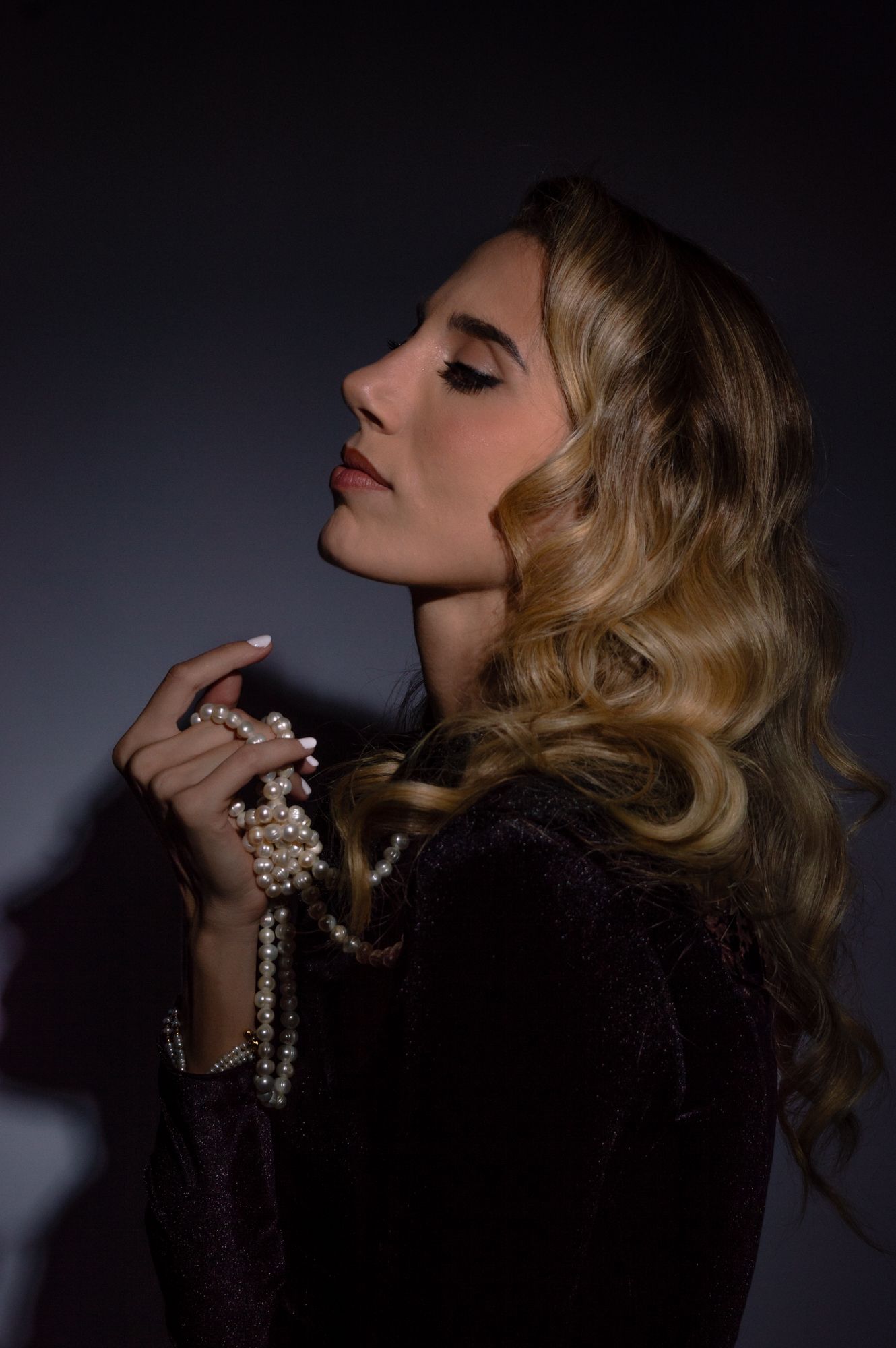 Glamorous blonde woman holding pearl necklace in a dark, moody studio portrait.