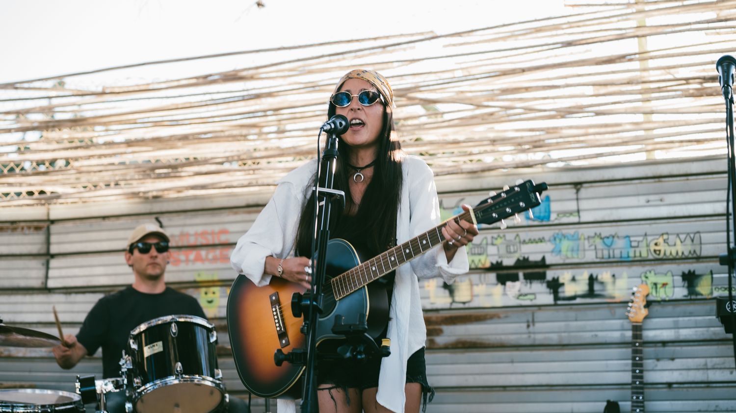 A female musician plays acoustic guitar and sings into a microphone on an outdoor stage with a drummer performing behind her.