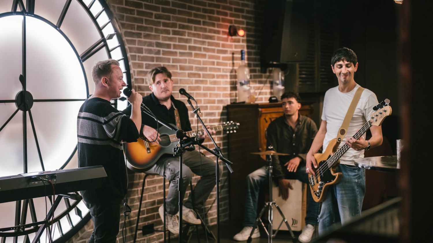 Four musicians perform acoustically in a brick-walled studio with a large clock backdrop.