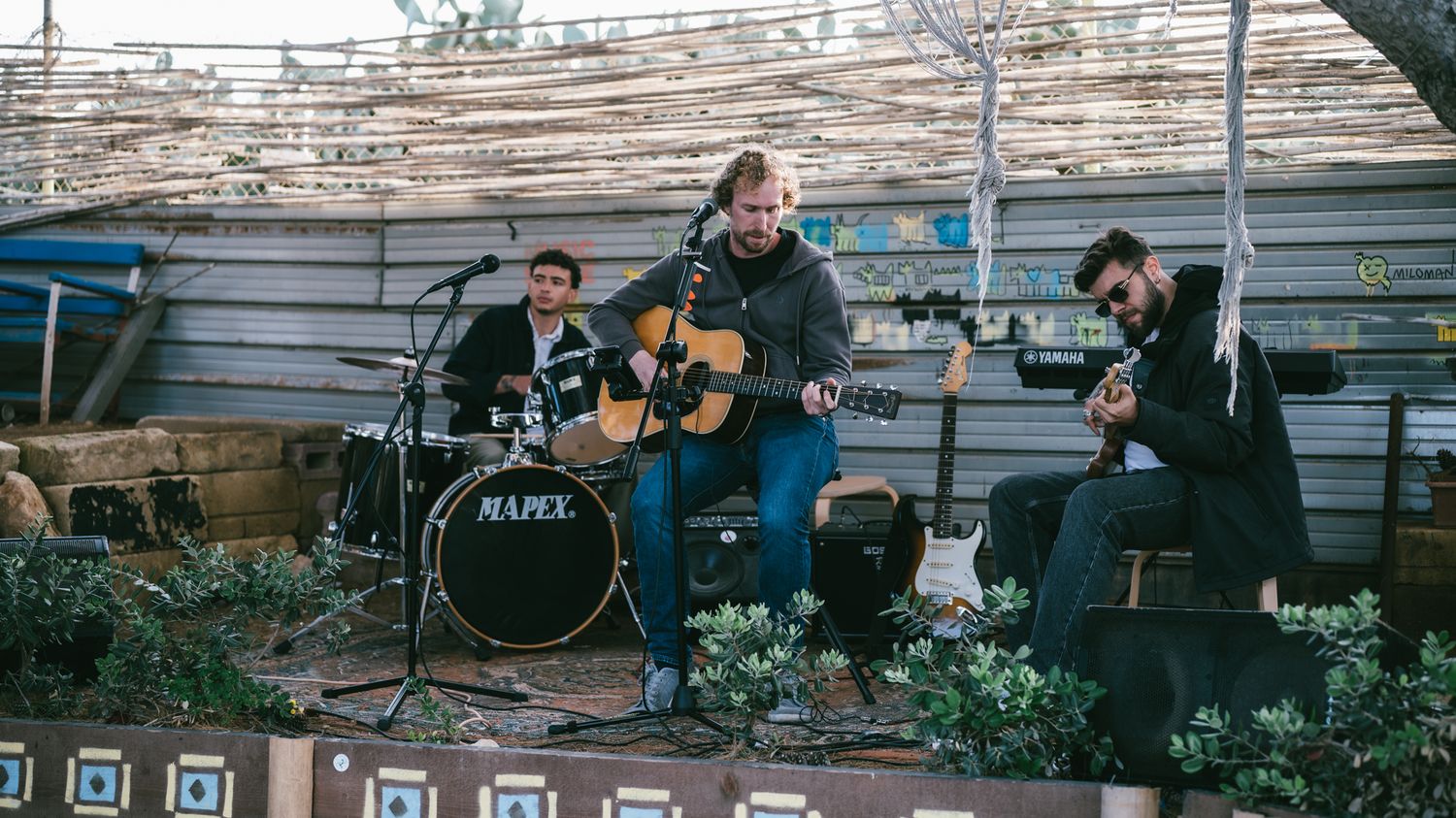 Three musicians perform outdoors, with a guitarist, drummer, and guitarist on a rustic stage surrounded by greenery.