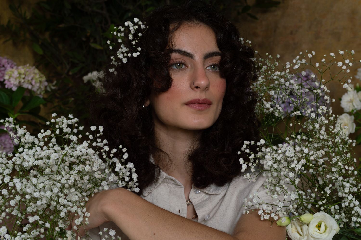 A young woman with curly dark hair poses serenely surrounded by white baby's breath and soft floral blooms.