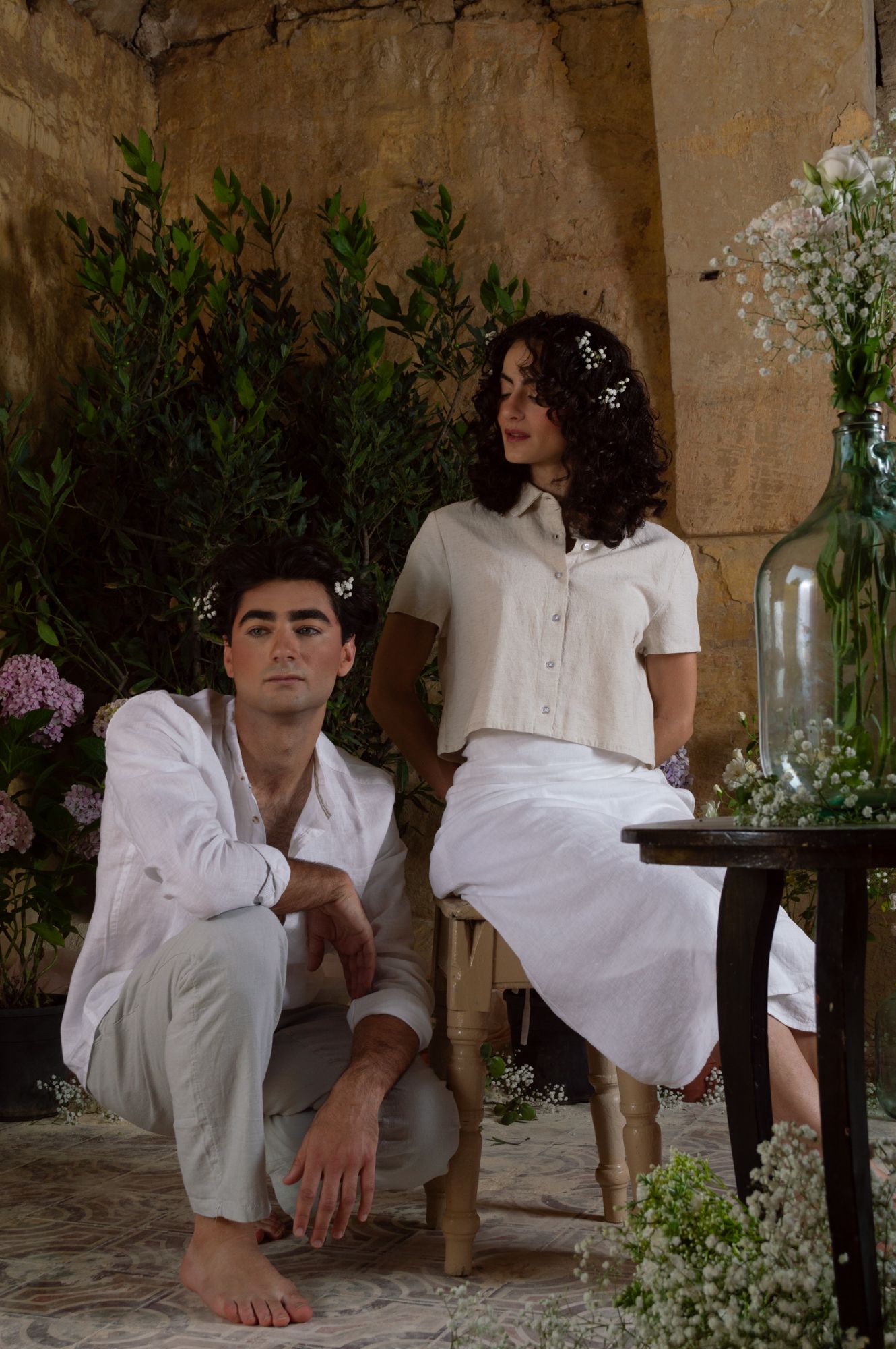 A young couple in white linen outfits pose casually among flowers and greenery in a rustic stone setting.