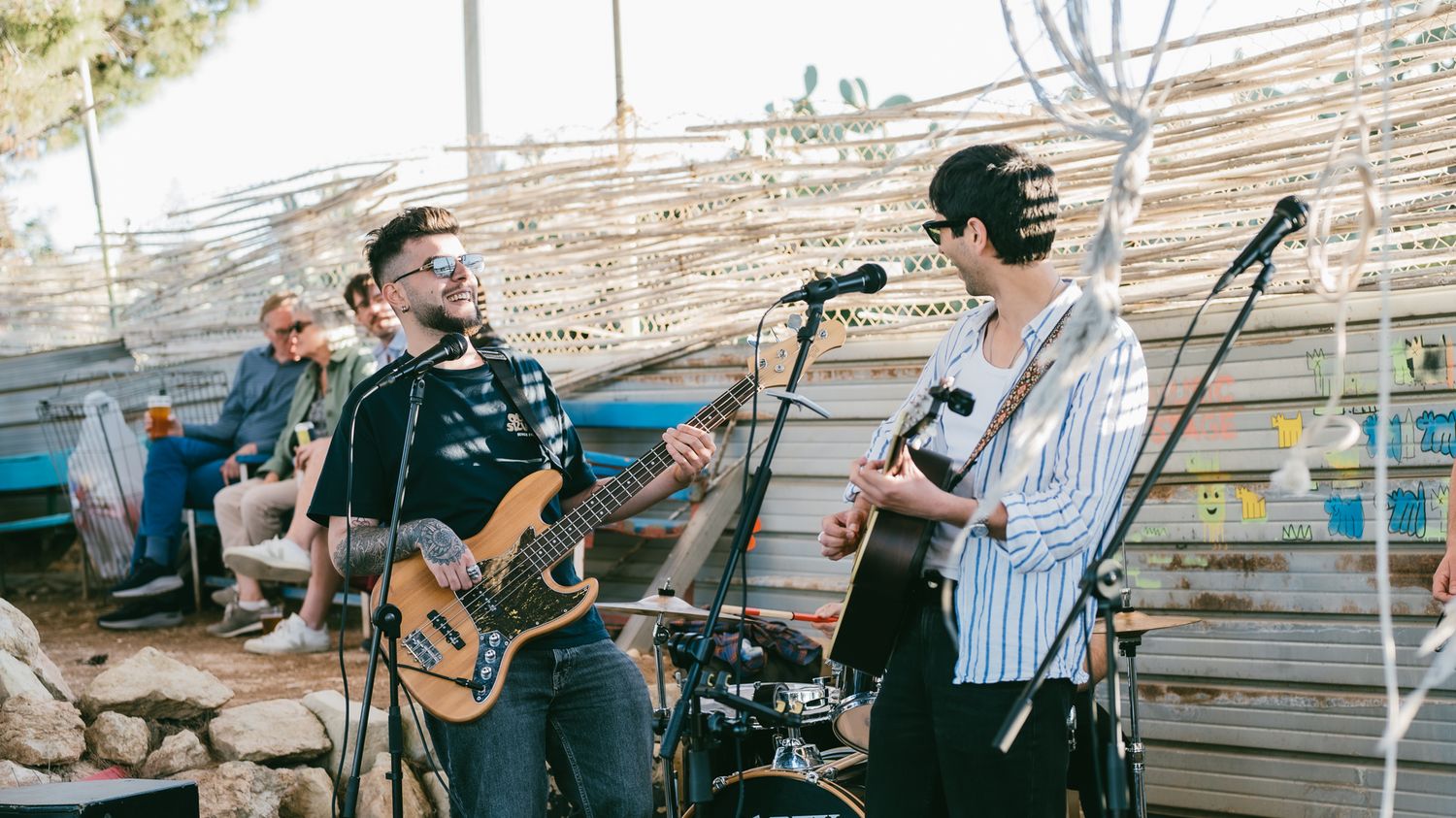 Two musicians playing guitar and bass on an outdoor stage at a sunny live music event.