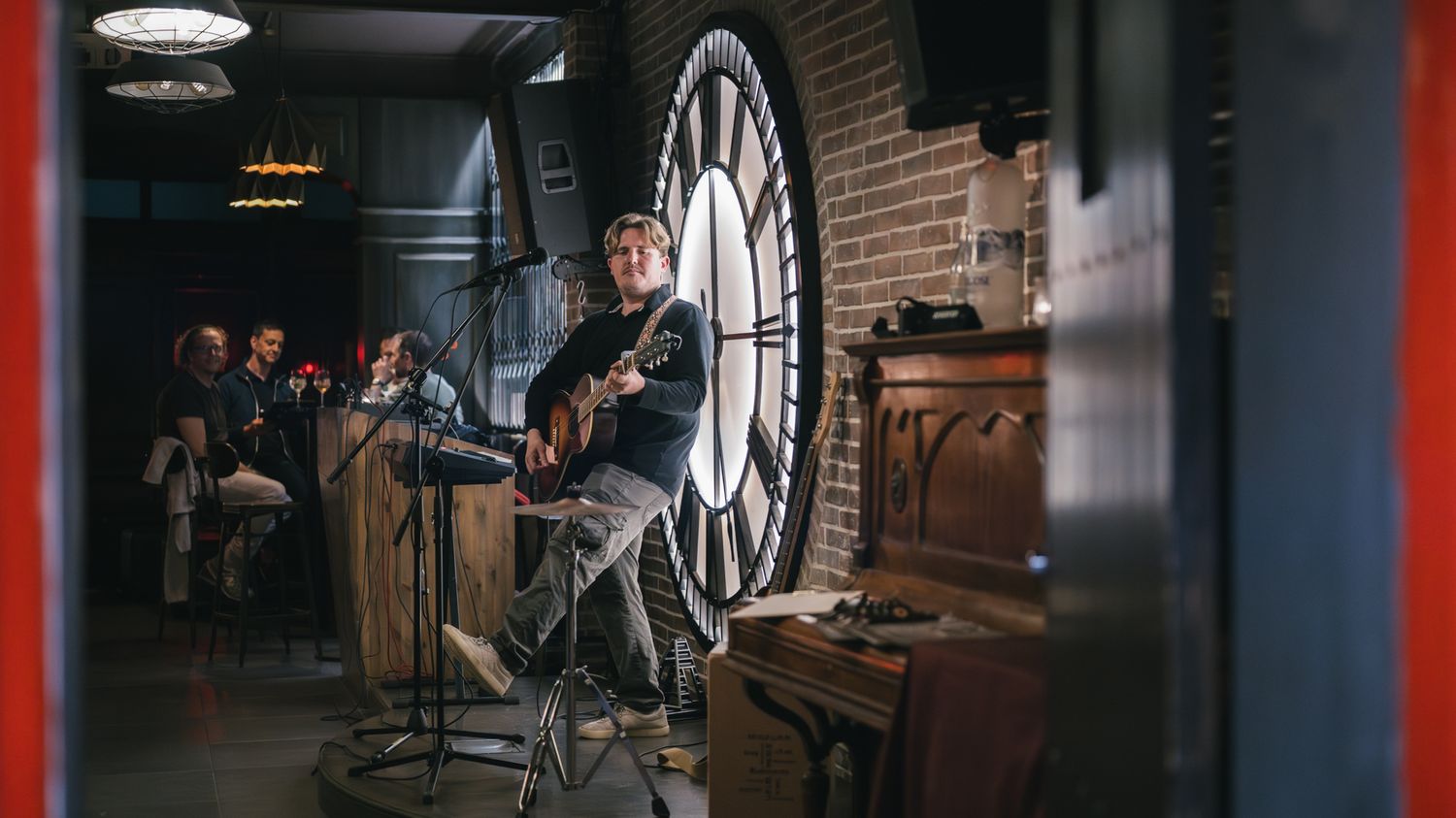 Musician playing acoustic guitar at a live indoor performance near a large clock and brick wall.