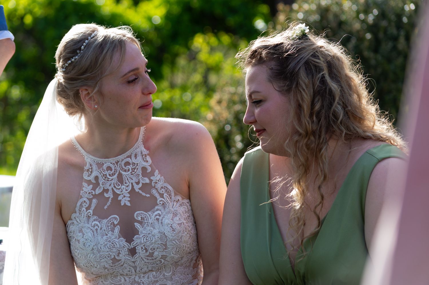 Bride in lace wedding dress and bridesmaid in green sharing an emotional moment at outdoor wedding.