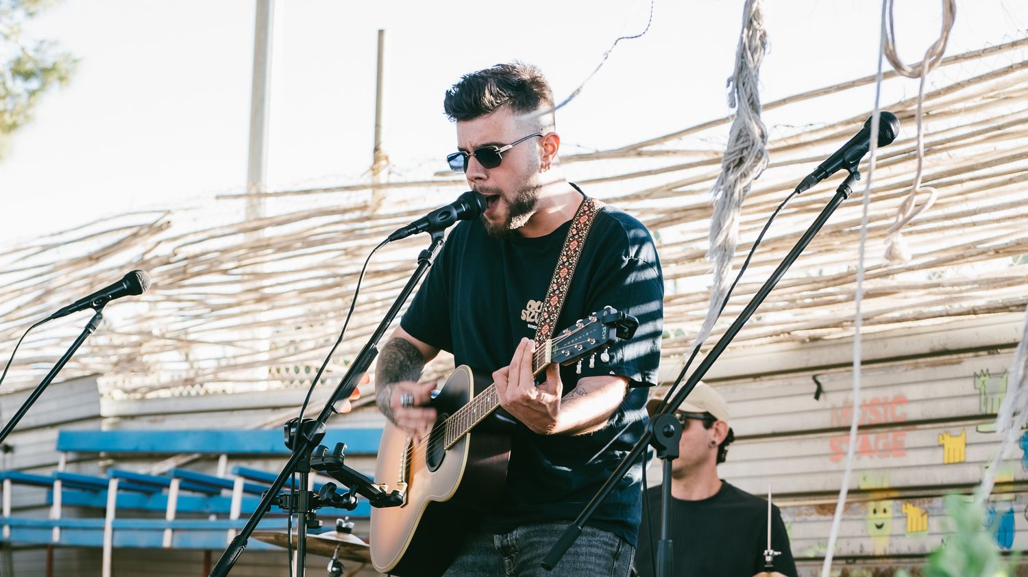 Male guitarist performing acoustic set outdoors at festival, wearing sunglasses and black shirt near microphone stand.