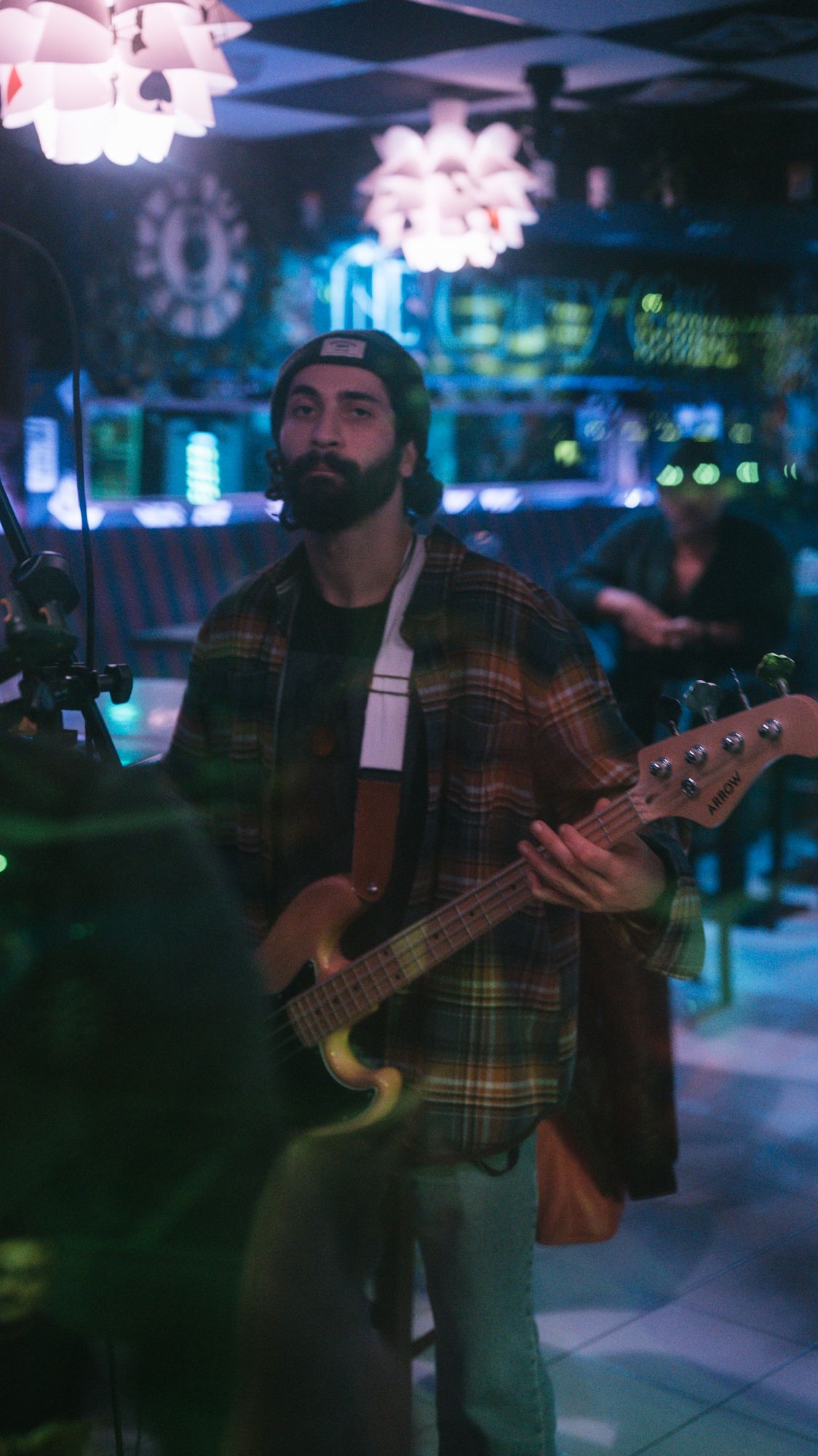 Bearded male bassist in a plaid shirt playing electric bass guitar at a colorful bar venue.