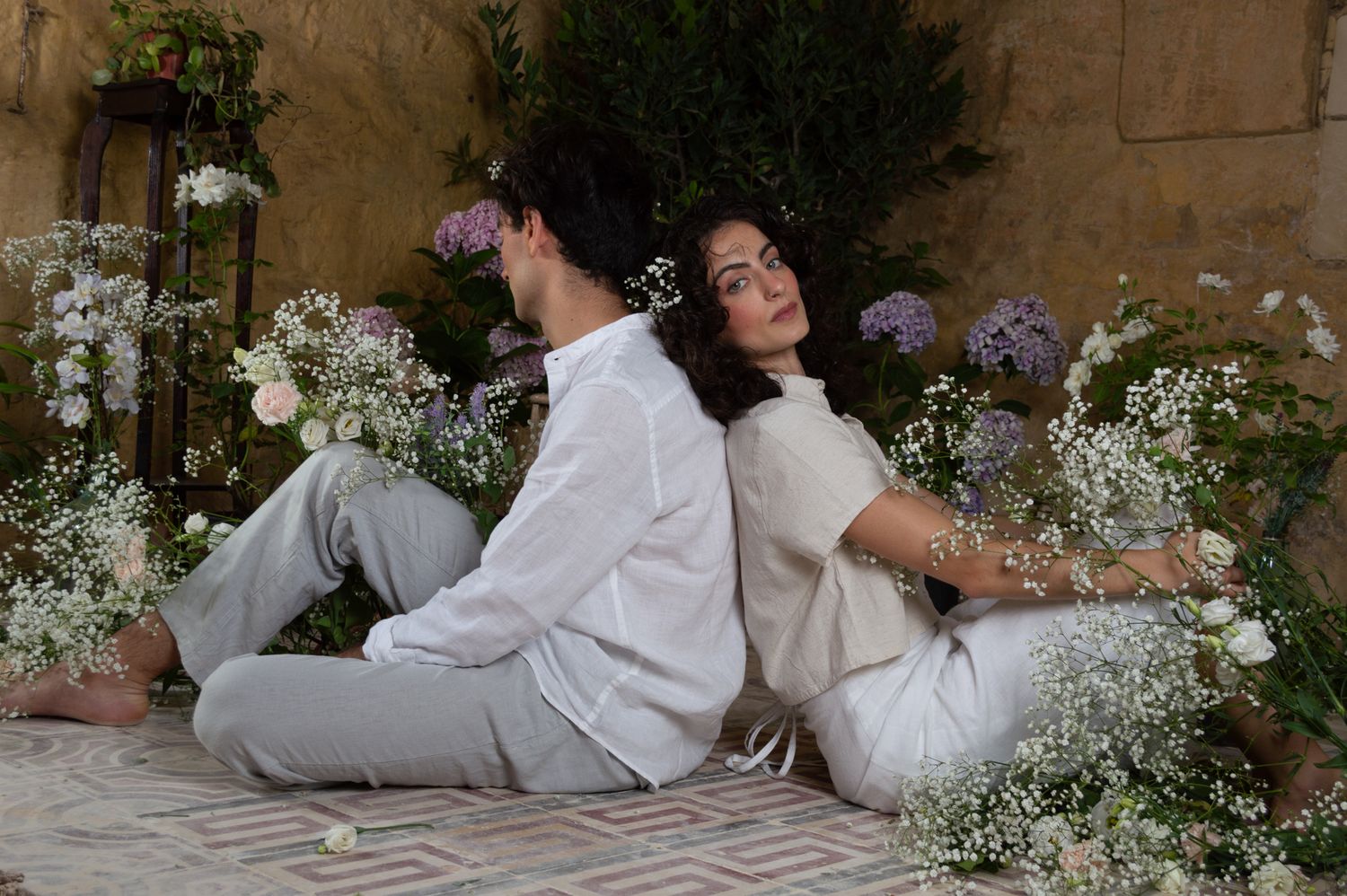 Couple in white linen sitting back-to-back surrounded by blooming flowers in a rustic courtyard.