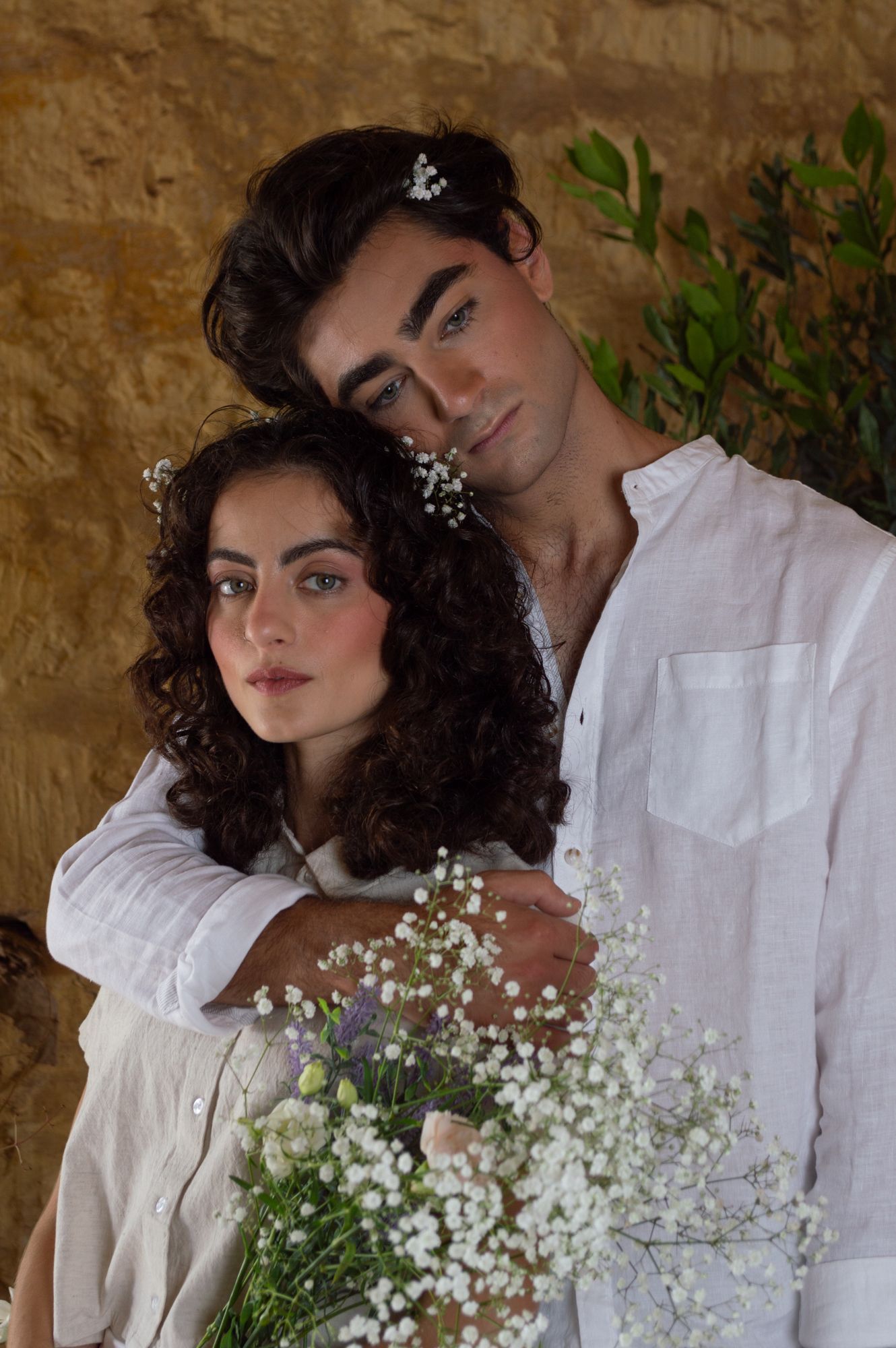Romantic couple in white linen embracing while holding a bouquet of baby's breath and wildflowers.
