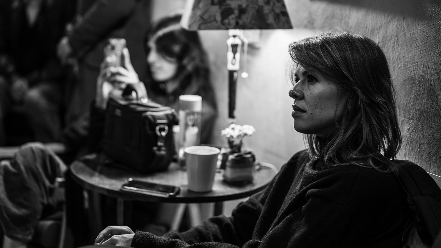 Black and white photo of a woman sitting alone in a cafe, holding a coffee cup, with a vintage camera on the table.