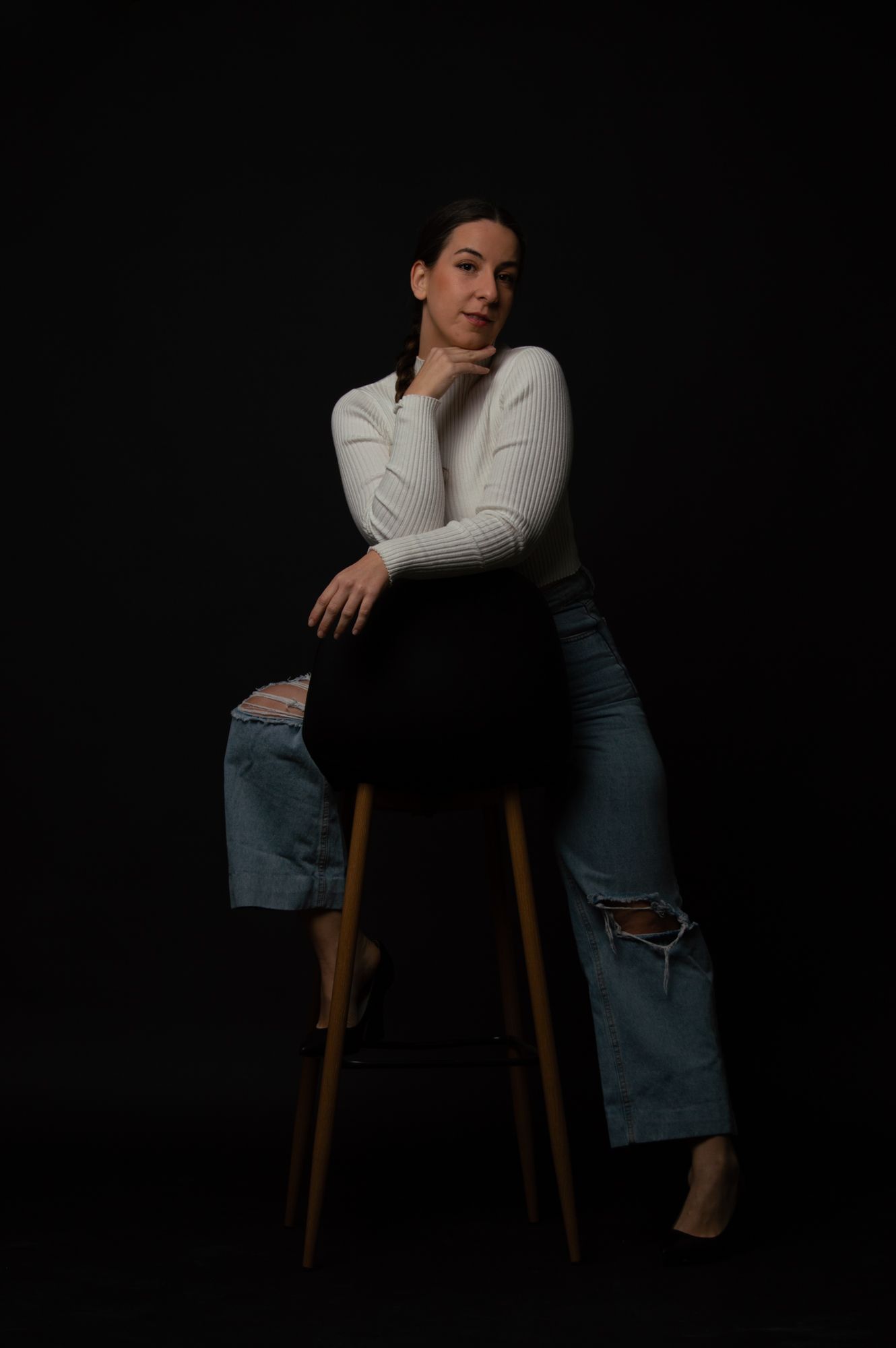 Woman in white sweater and ripped jeans sitting on wooden stool.