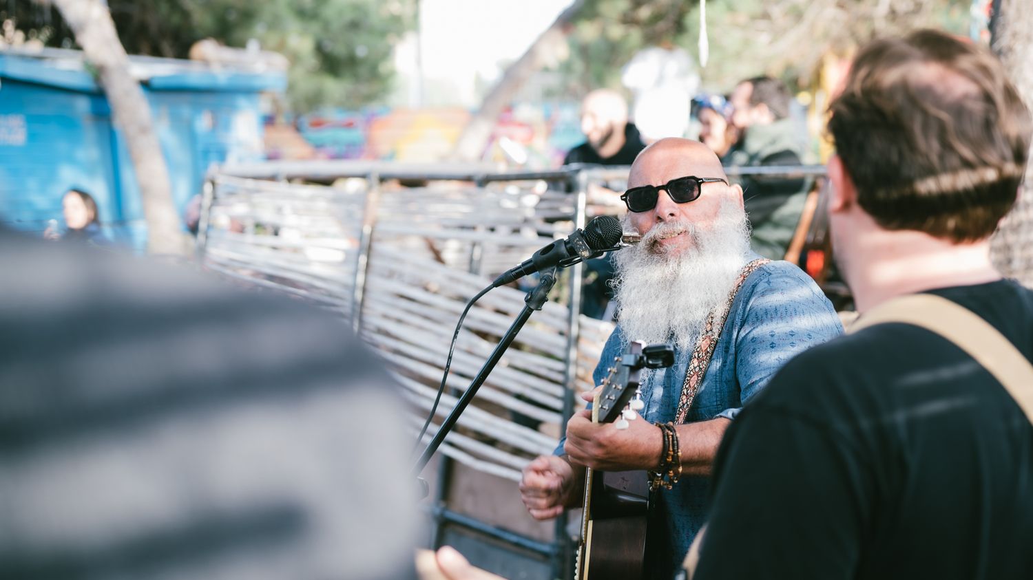 Elderly bearded musician with sunglasses performs acoustic guitar at outdoor festival near vintage truck.