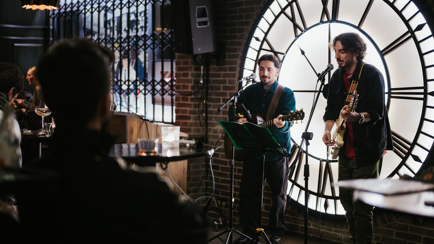 Two musicians perform acoustic guitars in front of a large clock face in a cozy brick-walled venue.