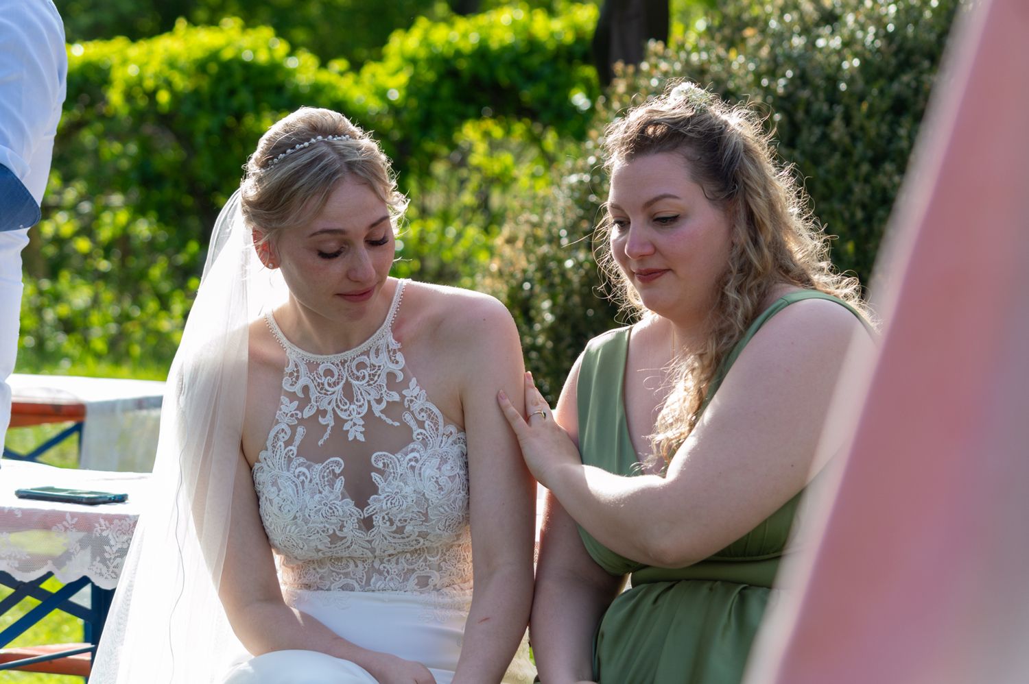 Bride in white lace gown and veil sits outdoors as bridesmaid in green dress offers comfort at wedding.