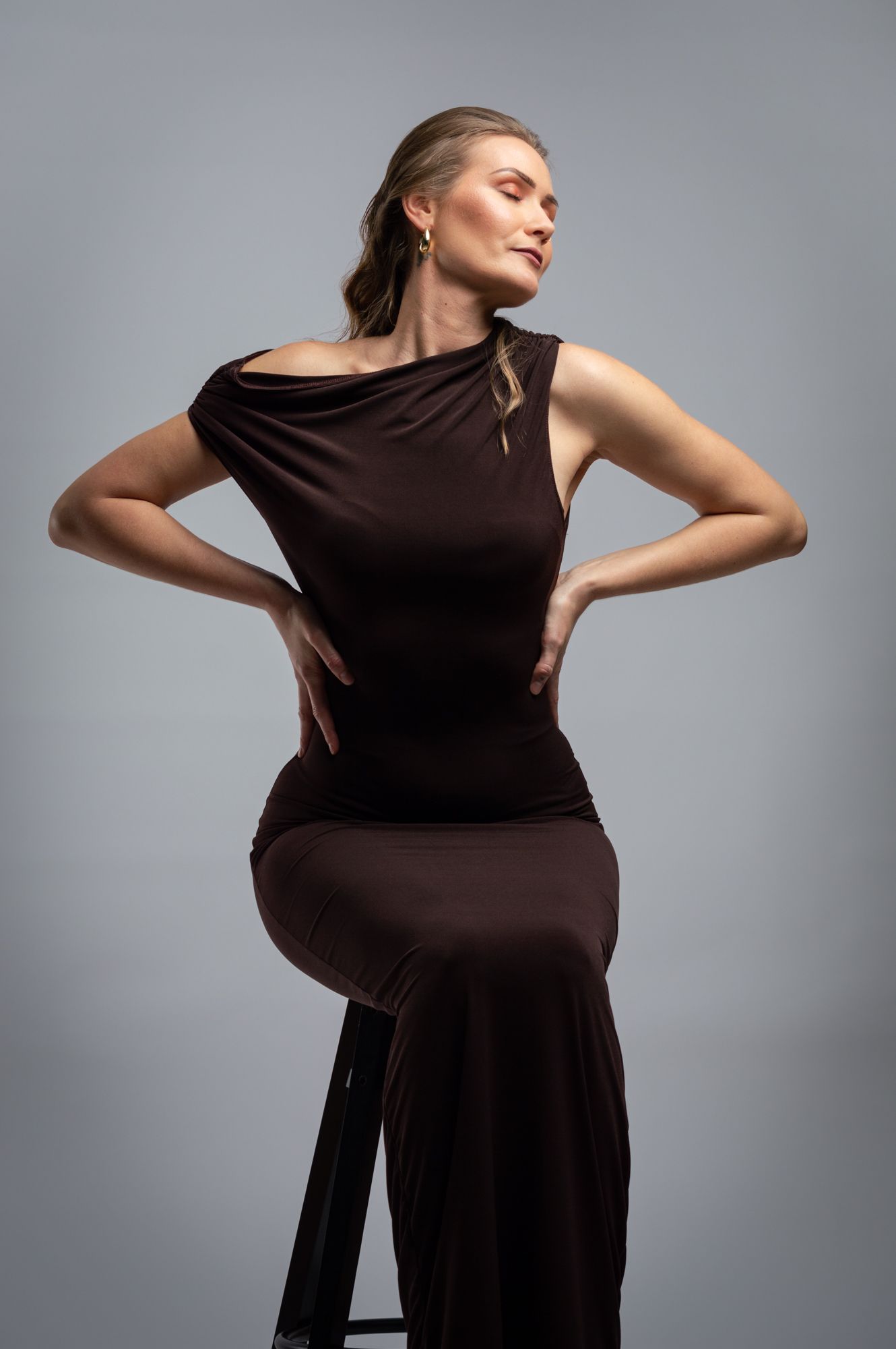 Woman in a sleek brown dress posing with hands on hips on a stool in a studio setting.
