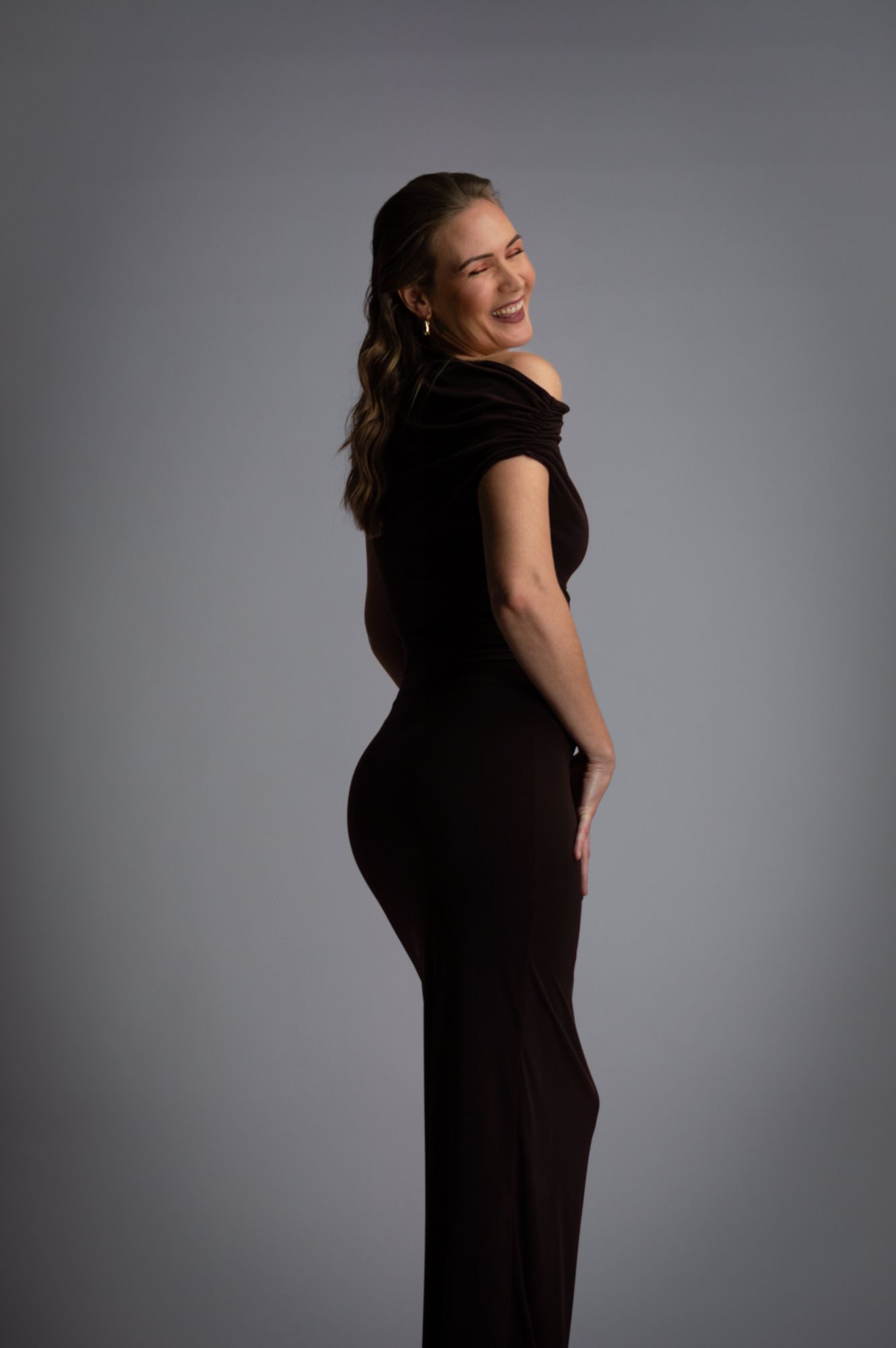 Laughing woman in black off-shoulder jumpsuit posing sideways in studio.