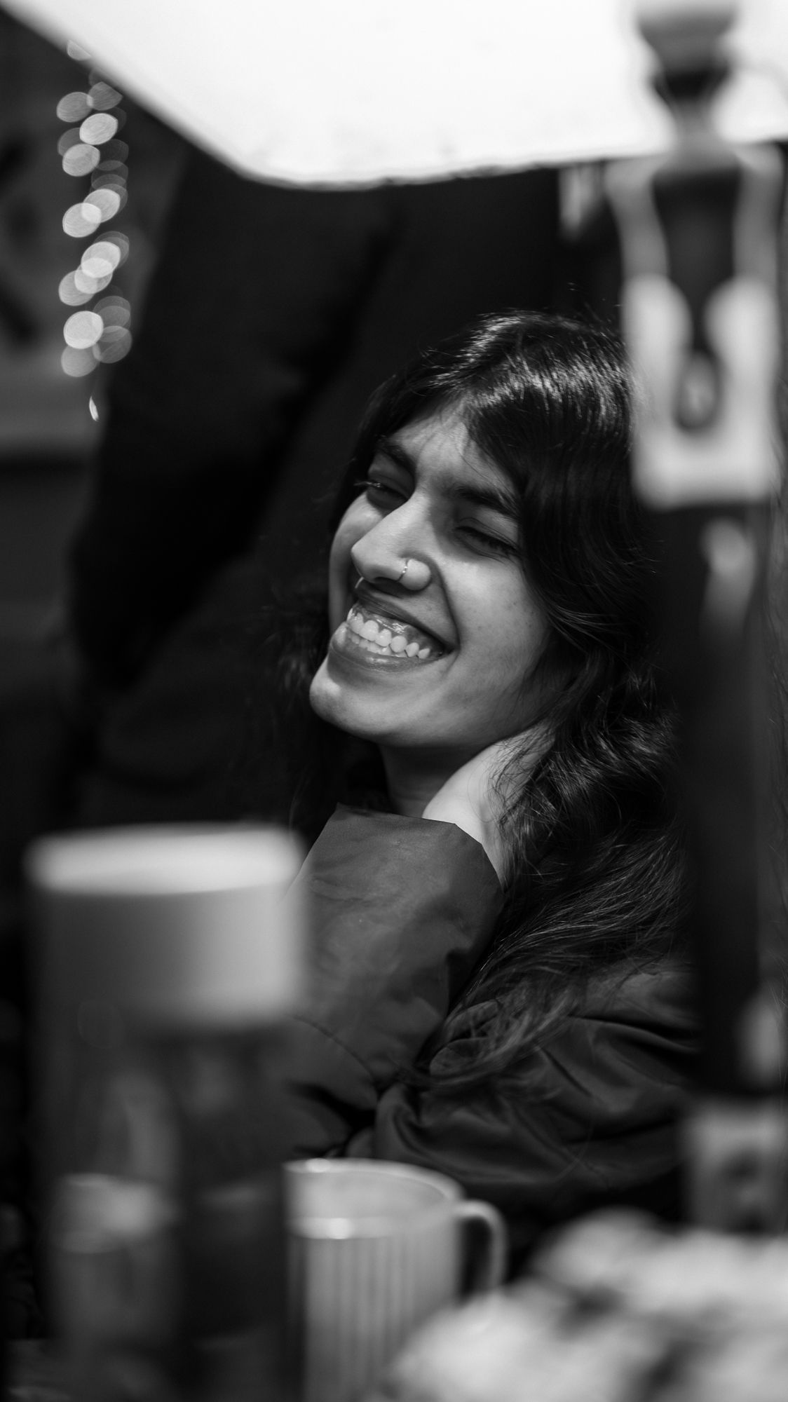Black and white portrait of a young woman laughing joyfully, seated near mugs in a warmly lit indoor setting.