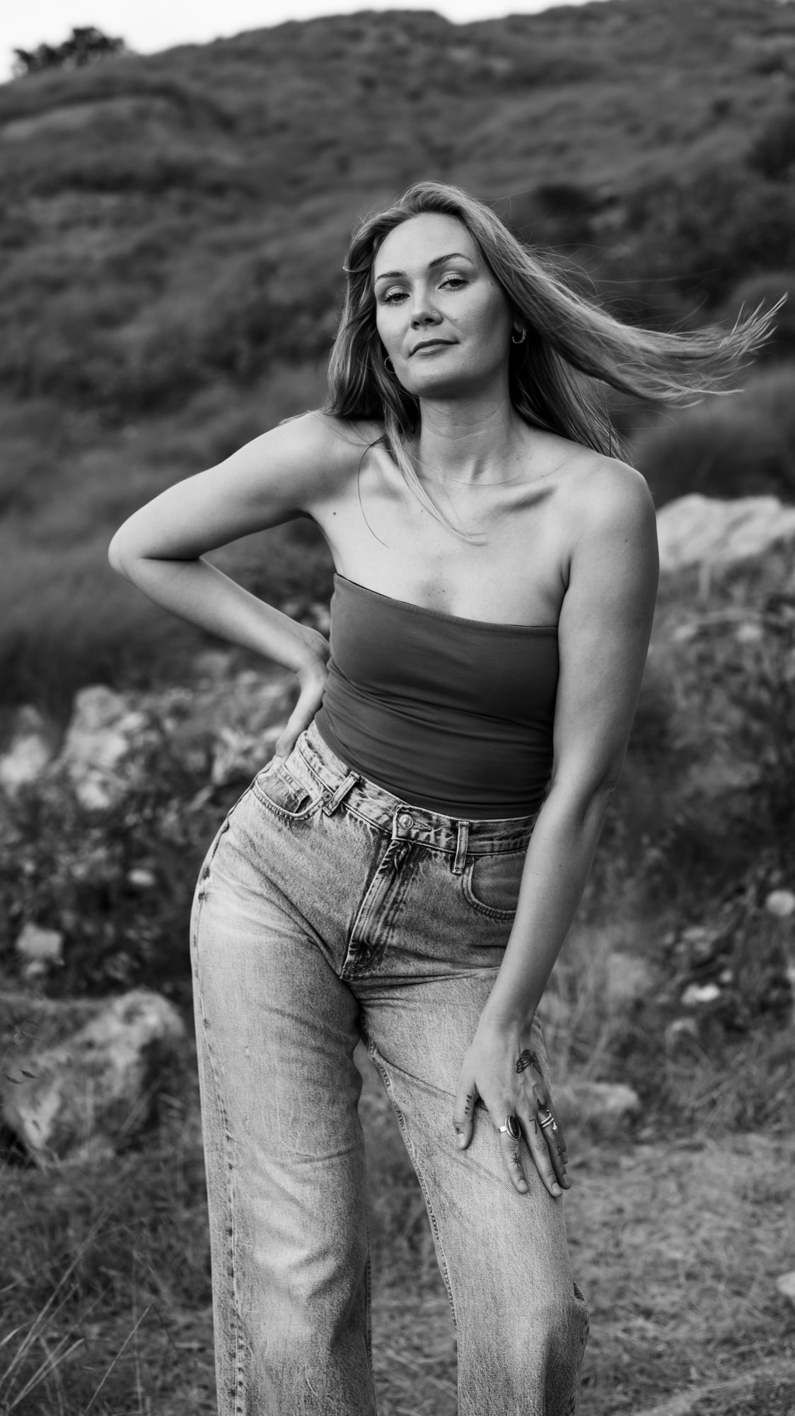 Black and white photo of a woman in a tube top and jeans posing outdoors with wind-blown hair.