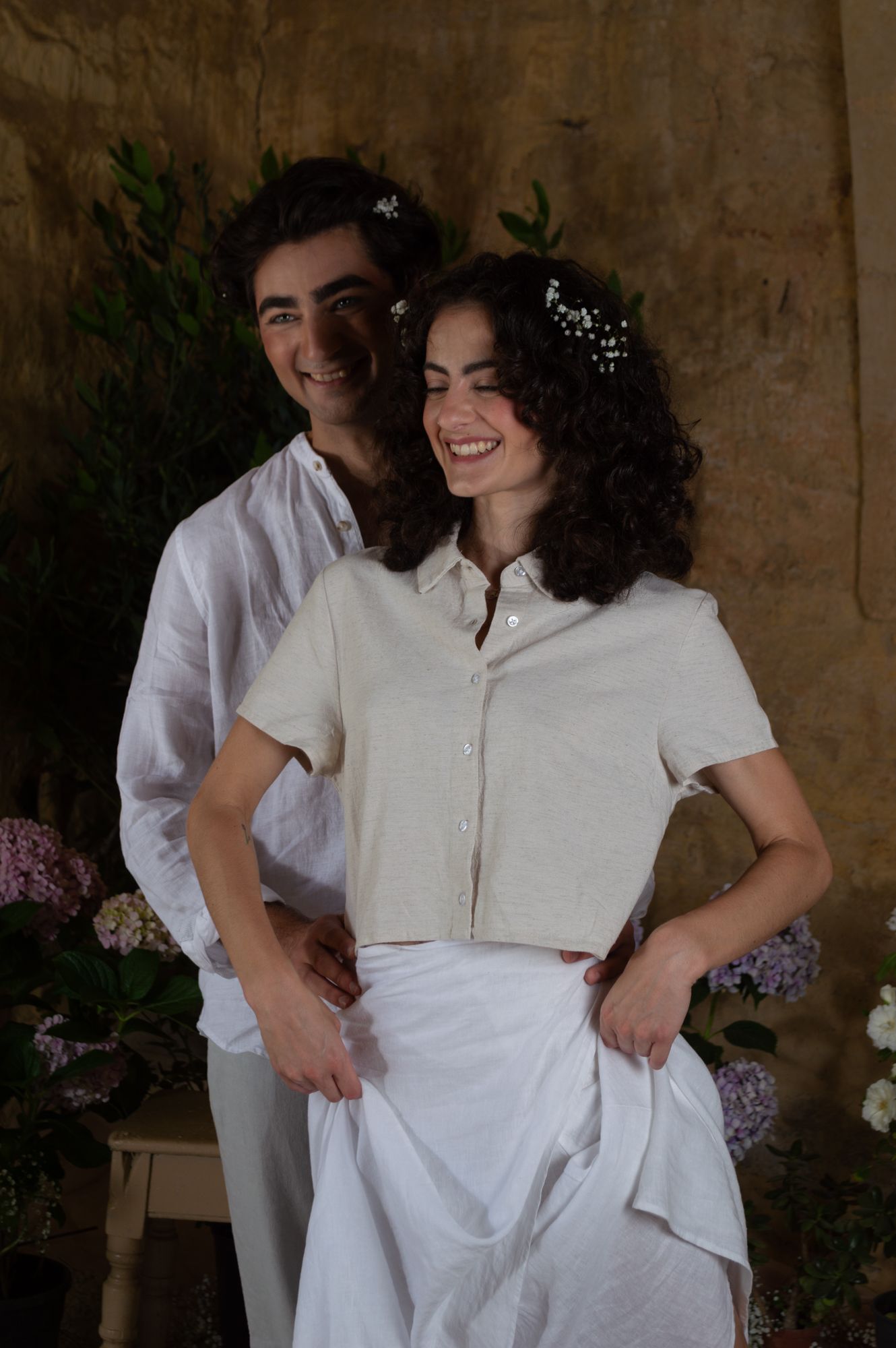 Joyful couple in white linen outfits laughing together among colorful hydrangeas in a rustic setting.