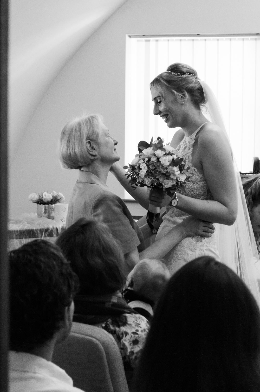 A bride in a white gown holds flowers while sharing a tender moment with an elderly guest at a wedding ceremony.