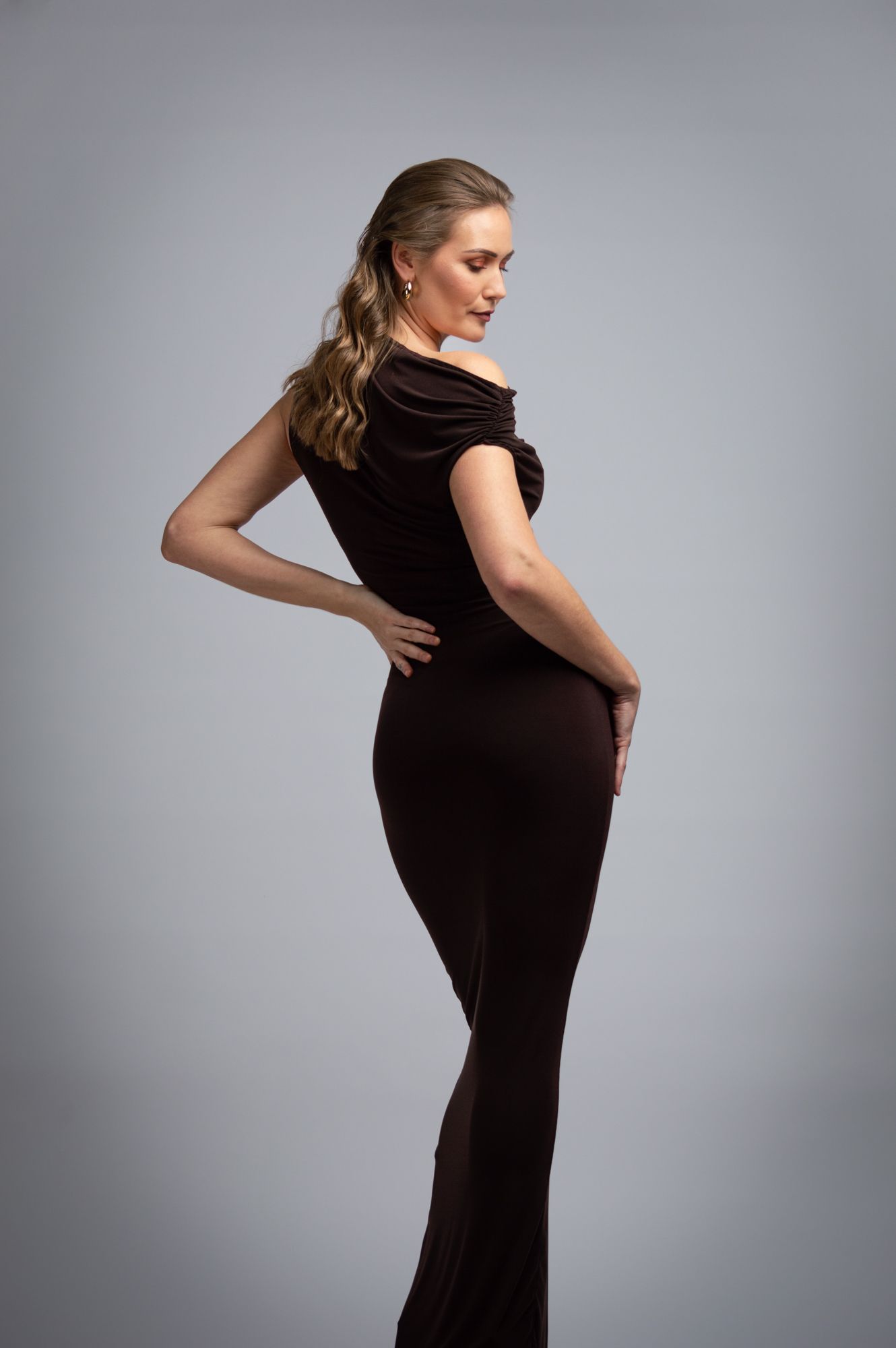Elegant woman in fitted dark brown off-shoulder gown posing in studio.