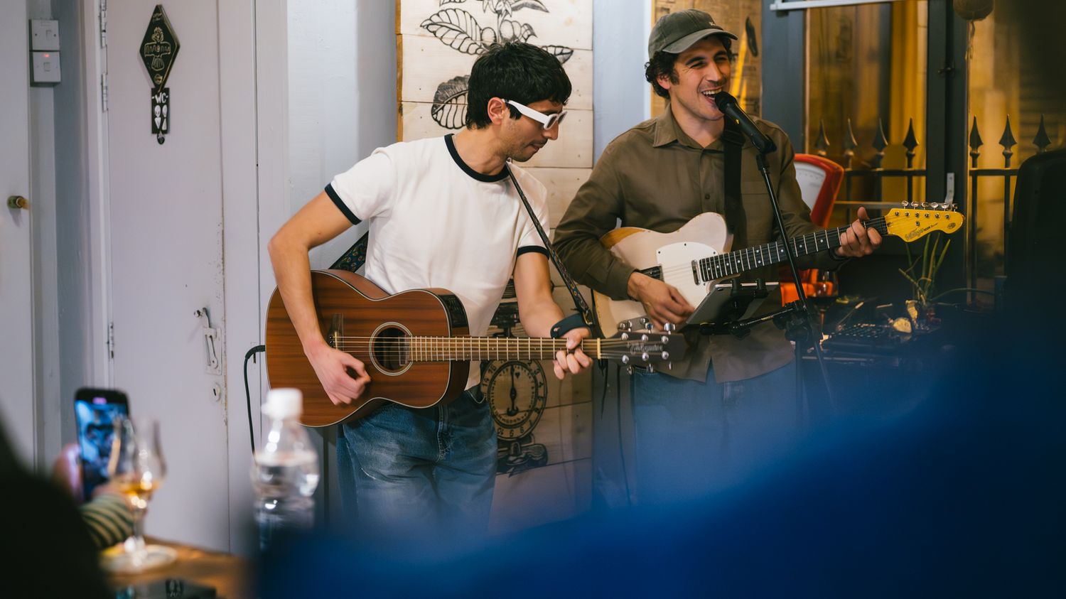 Two male guitarists performing acoustic and electric guitars at small indoor venue.