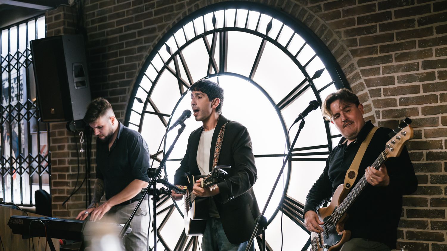 Three-piece band performing live in front of a large Roman numeral clock on a brick wall.
