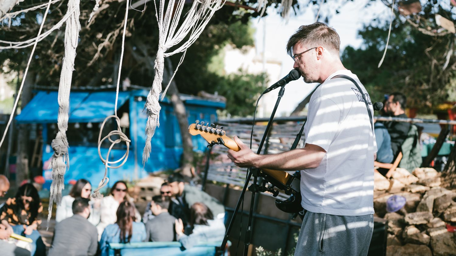 Musician playing electric guitar and singing at an outdoor event with guests dining under decorative hanging ribbons.