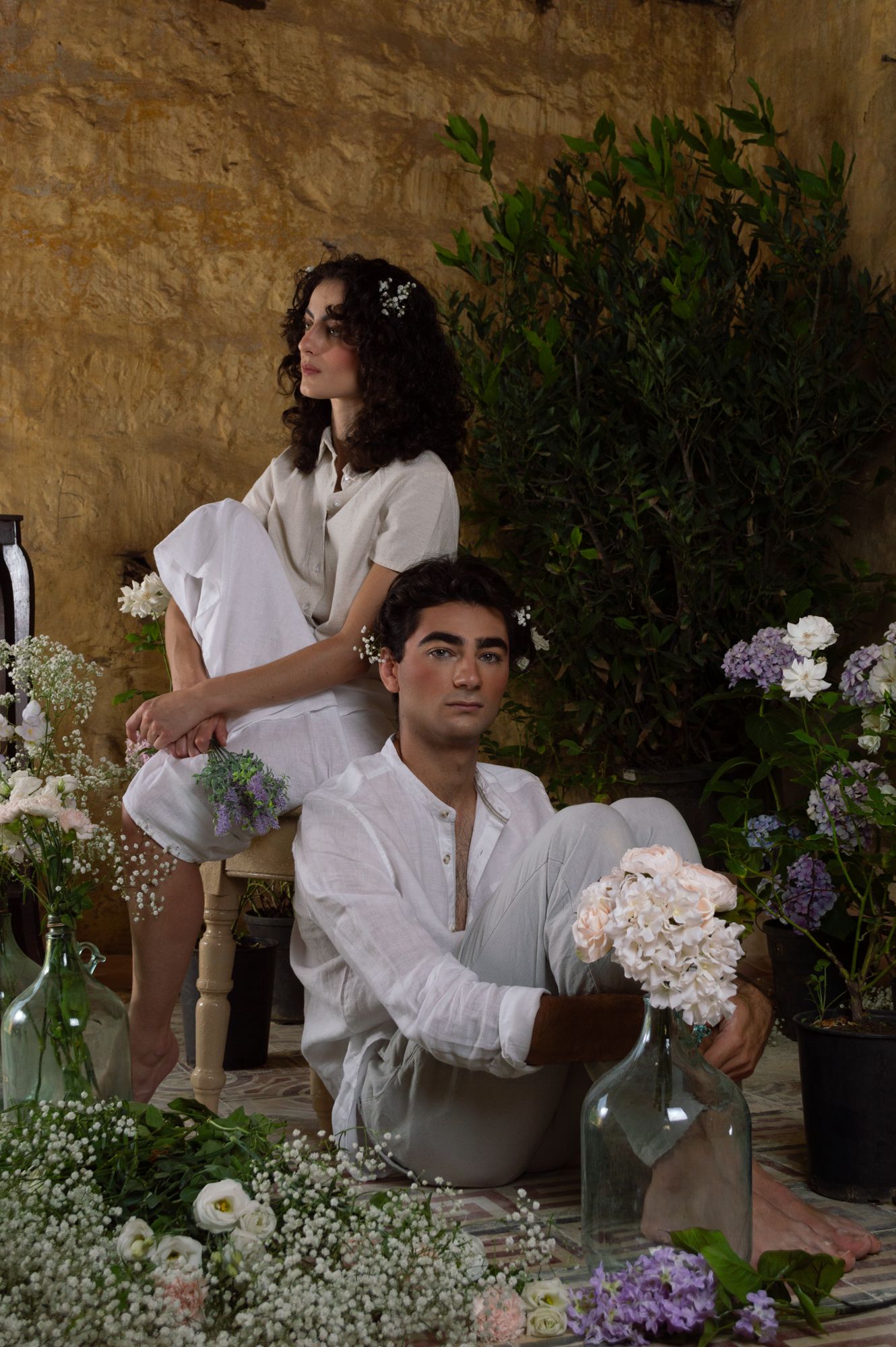 Couple in white linen outfits surrounded by fresh flowers and greenery in a rustic stone setting.